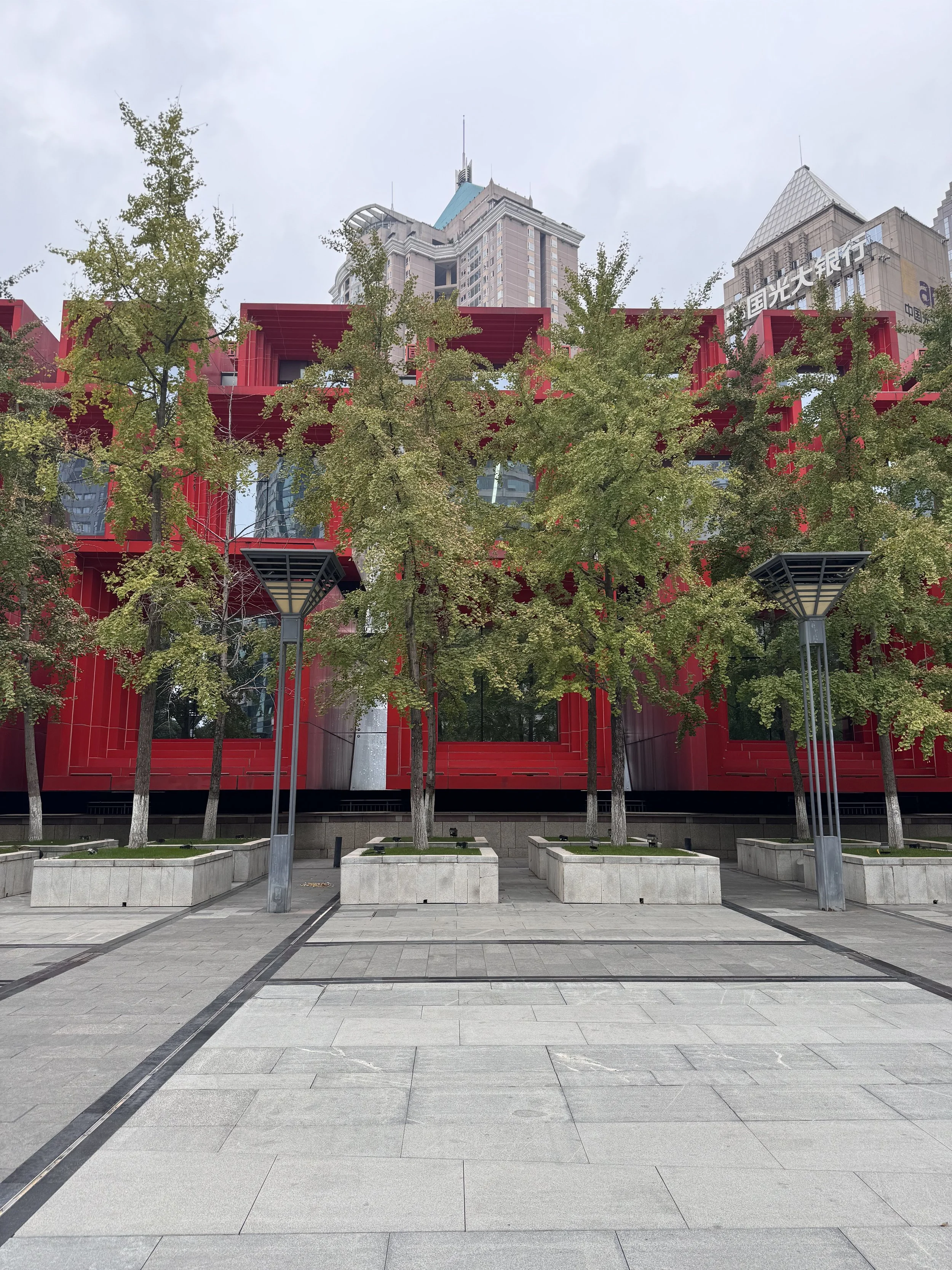 Urban plaza with gray stone pavement, small rectangular planters with trees, modern streetlights, and a red building with black windows and trees in front of it, with taller buildings in the background.