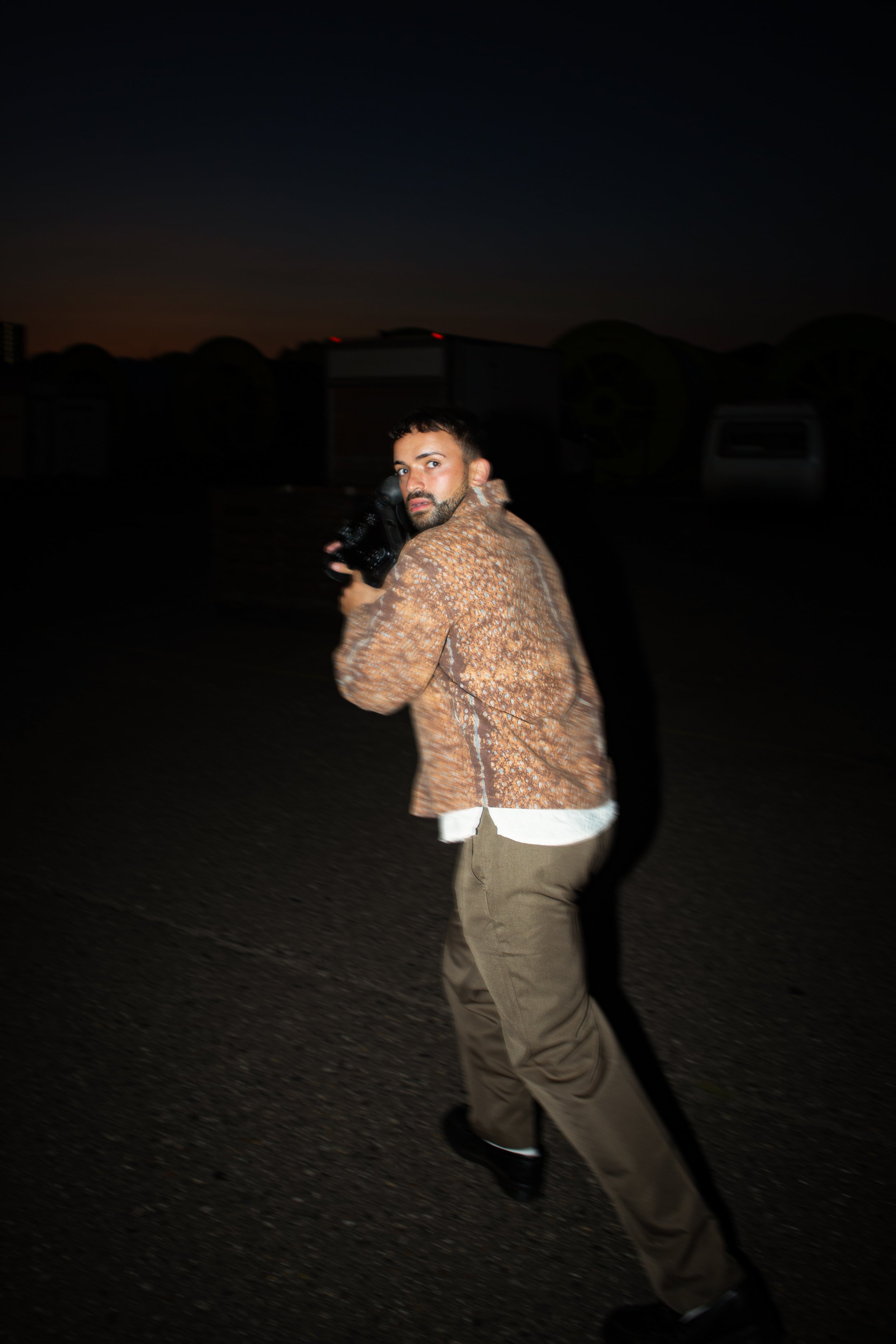 A man in a brown patterned jacket and beige pants holding a camera, looking over his shoulder at night on a dark street.
