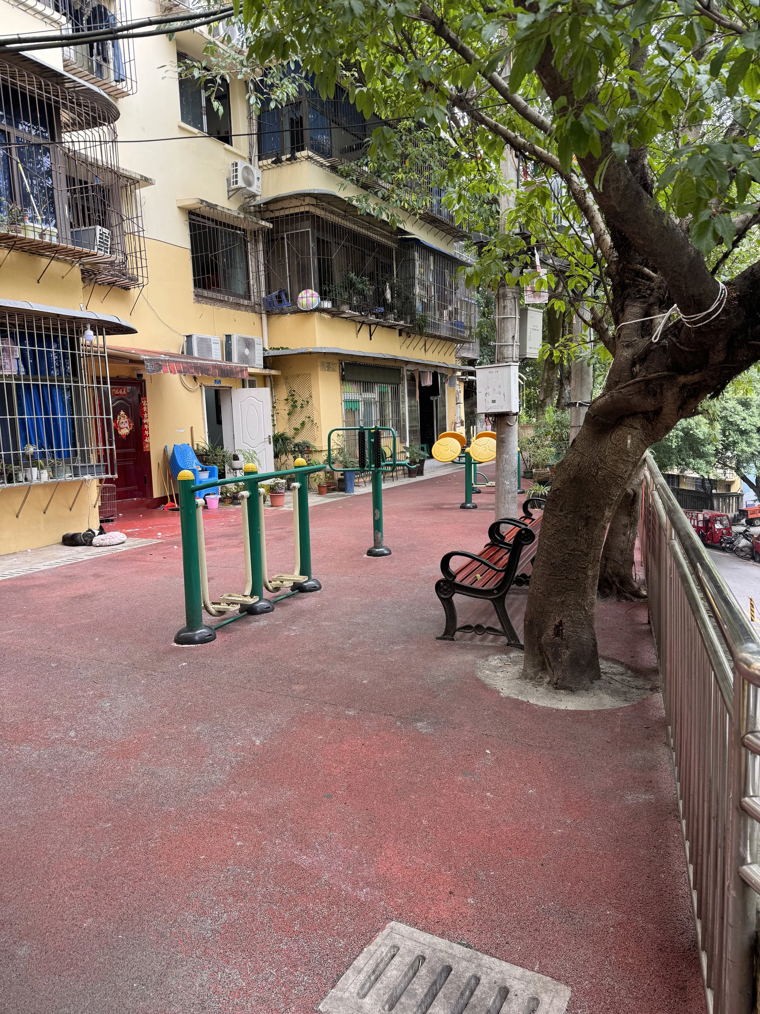 Outdoor playground area adjacent to a residential building, featuring exercise equipment, benches under a tree, and apartment balconies with metal grilles.