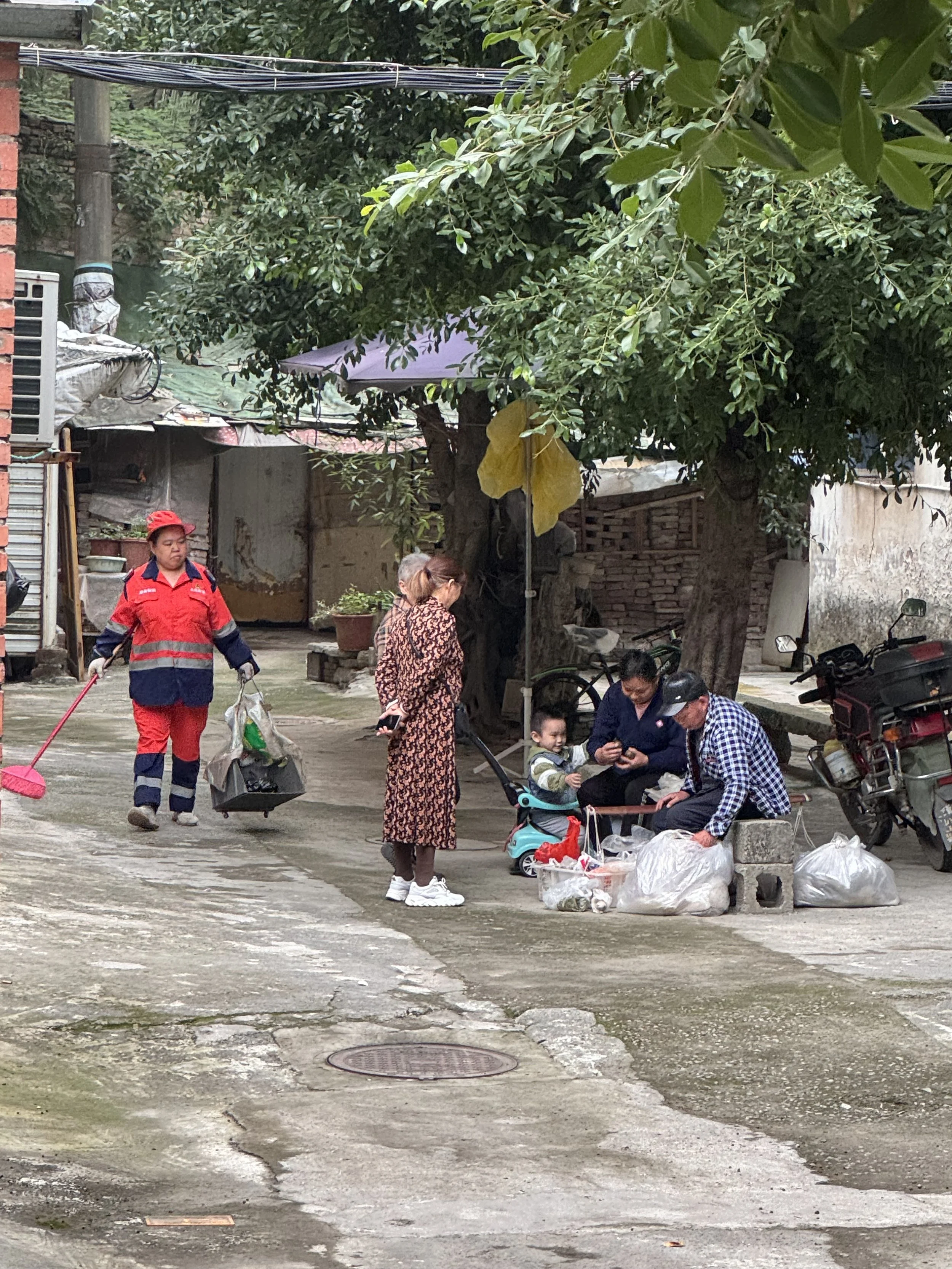 A group of people, including a child, gathered around on a street corner. There are three adults and one child. The adults are sitting and standing near plastic bags with items, possibly selling or trading. One woman in a red and navy uniform is sweeping the street. There are bikes and motorcycles parked nearby under trees providing shade.