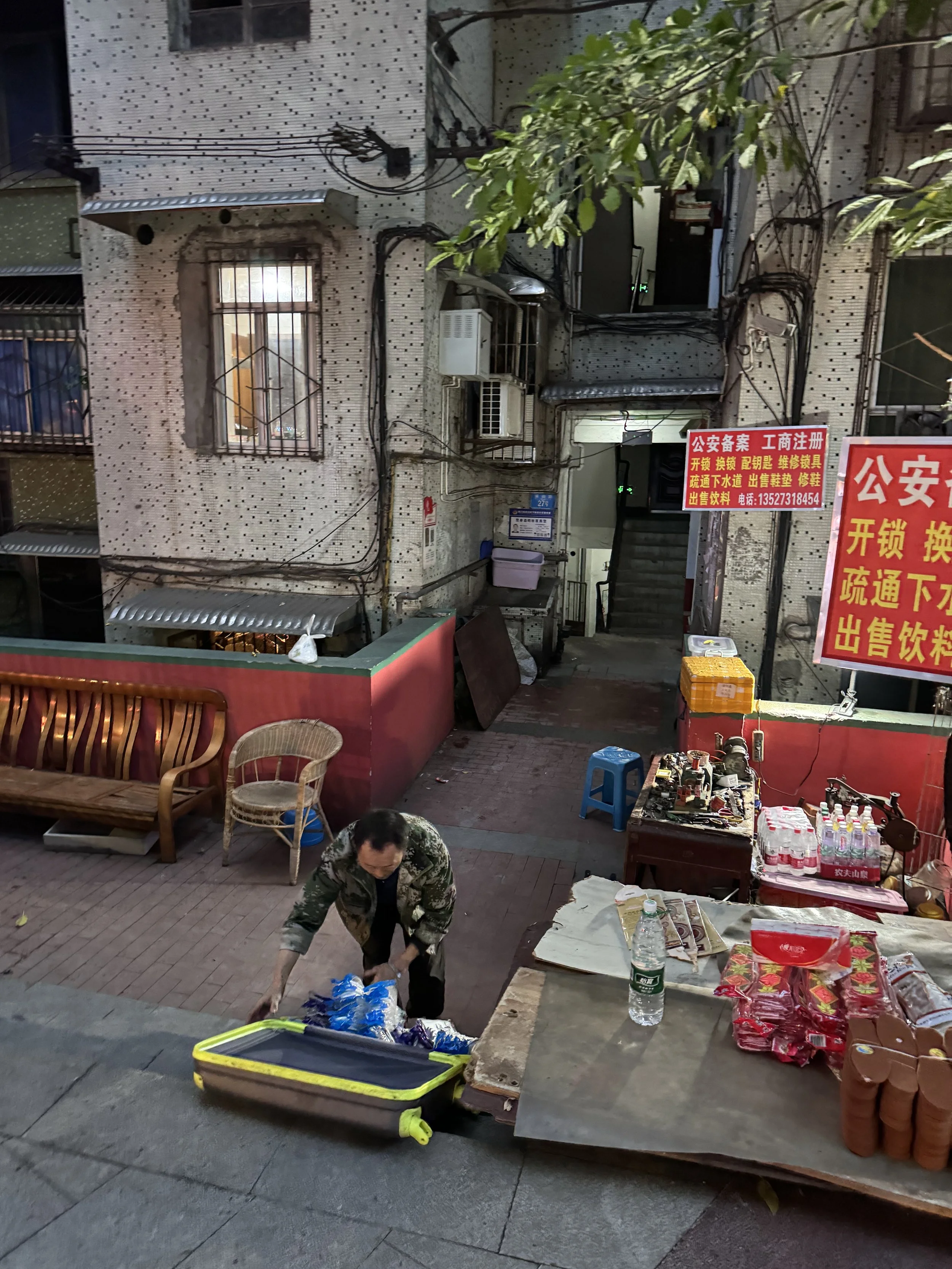 A man loading supplies into a yellow suitcase on a sidewalk in an urban area with a small outdoor market stand. There are chairs, a table with bottles and packages, and signs in Chinese in the background. The building behind has a textured wall, a window with bars, and various exposed wires.