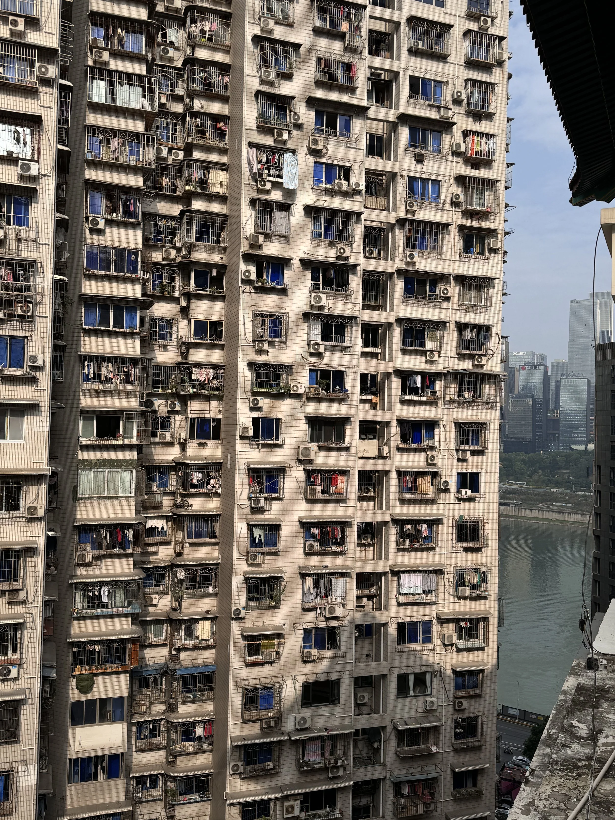 A tall residential apartment building with multiple balconies, each enclosed by metal bars, with various clothes and items hanging to dry, and numerous air conditioning units installed on the exterior.