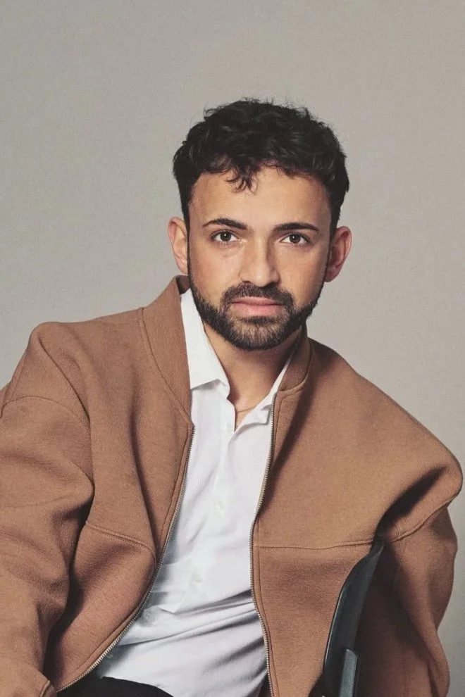A man with dark curly hair and a beard, wearing a brown jacket over a white shirt, sitting against a neutral background.