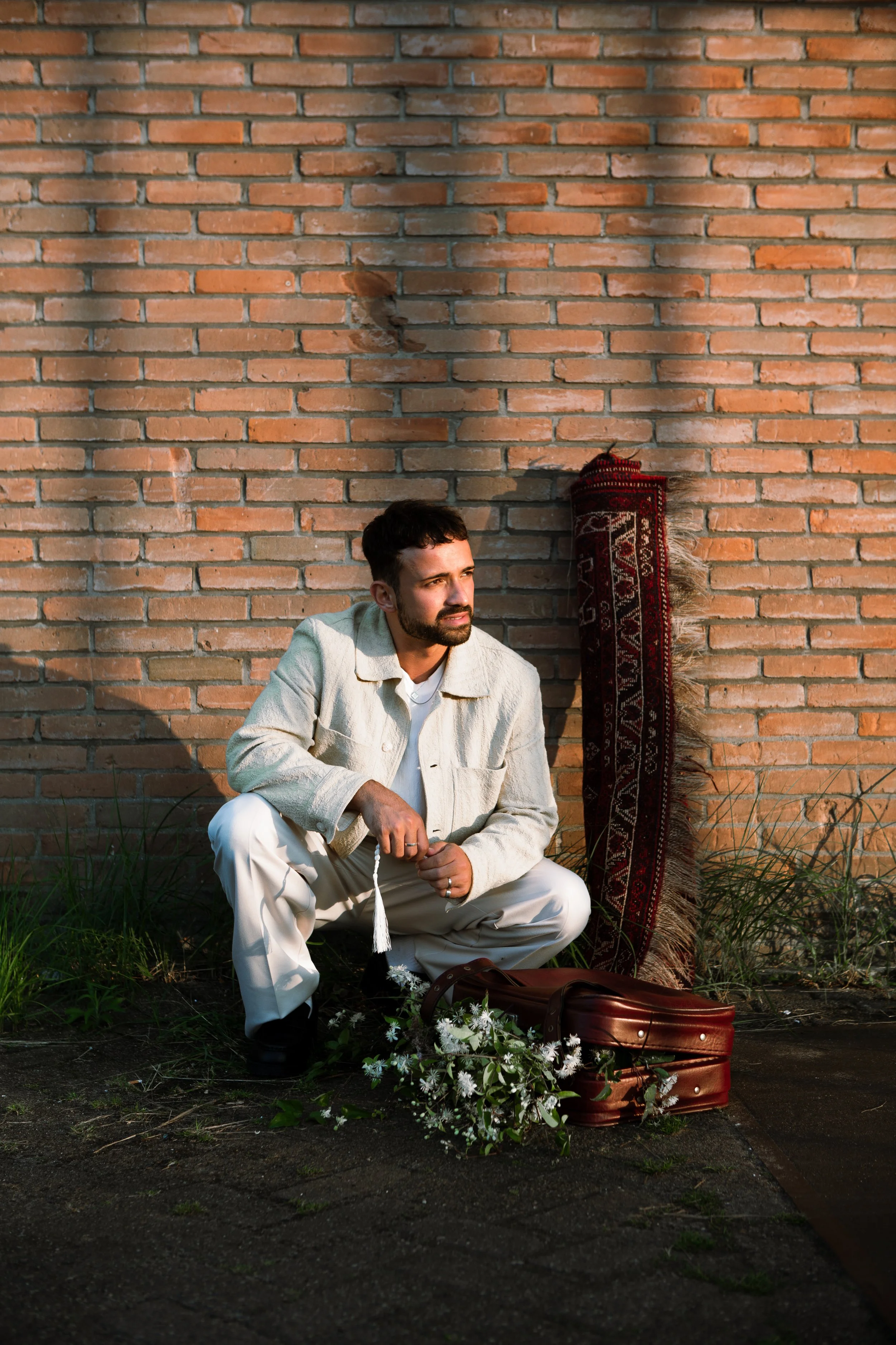 A man with dark hair and a beard is sitting on the ground next to a brick wall. He is dressed in a light-colored jacket and pants, holding a tassel in his right hand. There is a bouquet of white flowers and an open leather bag on the ground beside hi