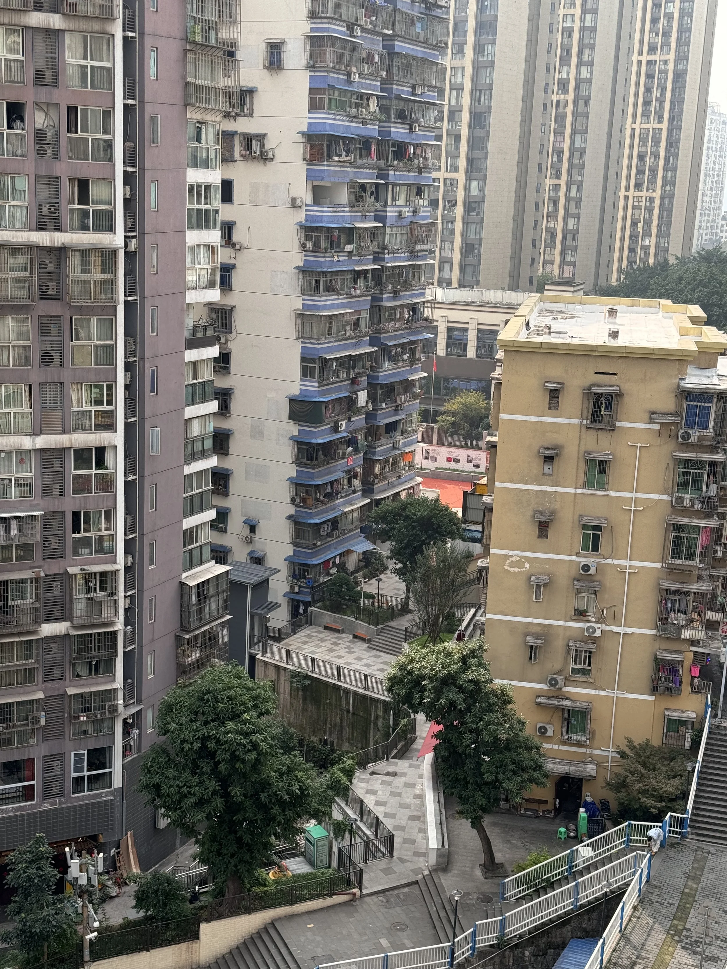 Cityscape view of multiple high-rise apartment buildings with balconies, trees, and a paved courtyard area with stairs and benches.
