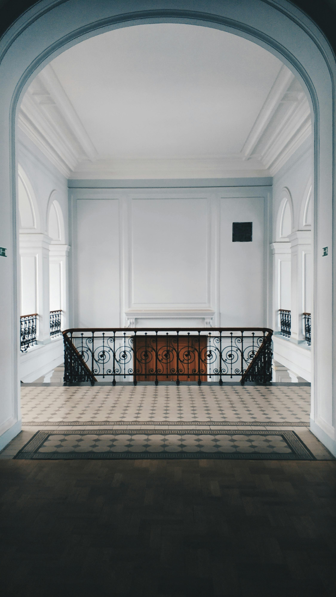 Interior view of a bright, white, elegant hallway with decorative moldings, arched windows, and a staircase with ornate black iron railings.