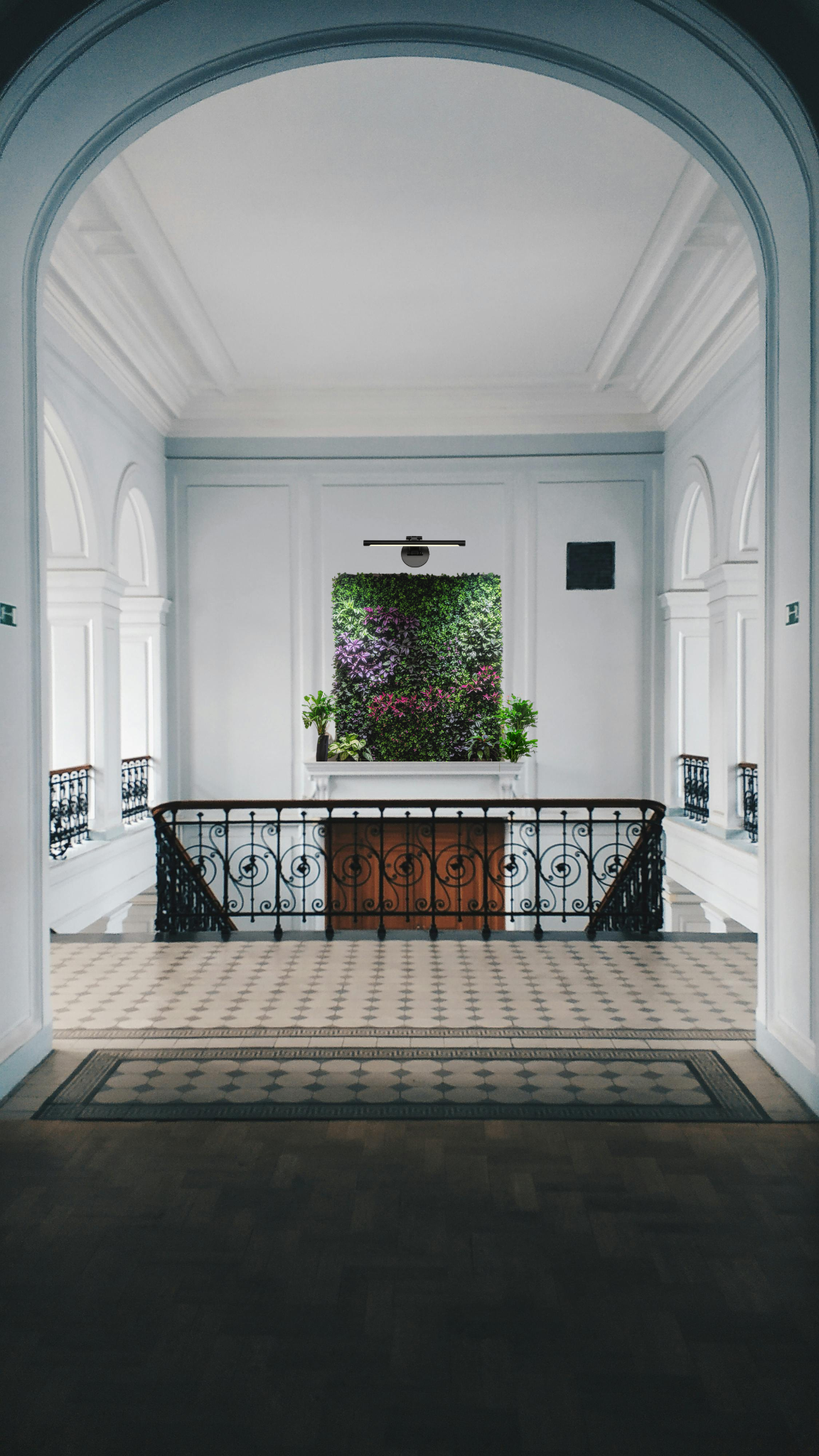Interior view of a white-walled corridor with decorative moldings, black wrought-iron railing, and a tiled floor, leading to a wall with a green and pink floral garden framed by a window.