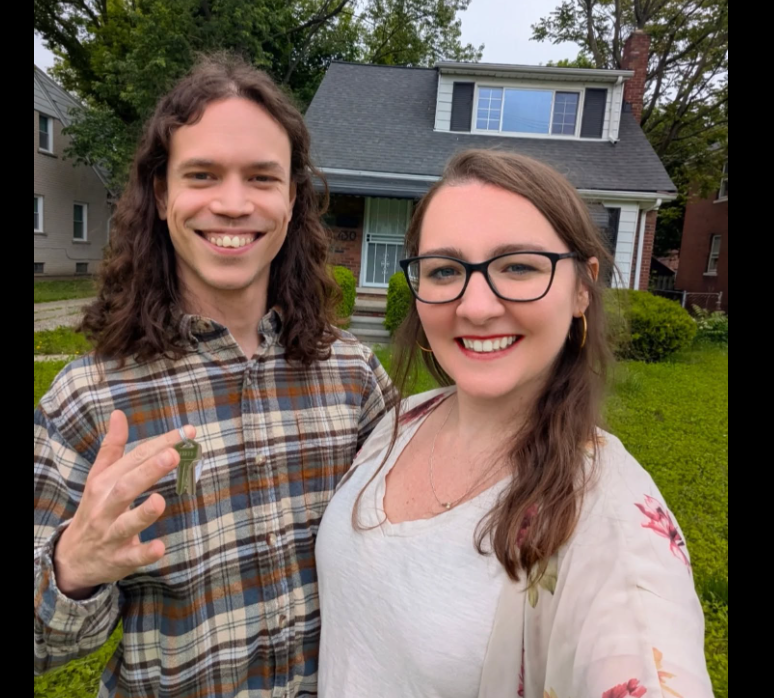 A smiling couple standing in front of a house with a gray roof and green lawn, taking a selfie. The man has long, wavy brown hair and is wearing a plaid shirt, holding a key in his hand. The woman has long brown hair, glasses, and is wearing a white shirt with floral patterns.