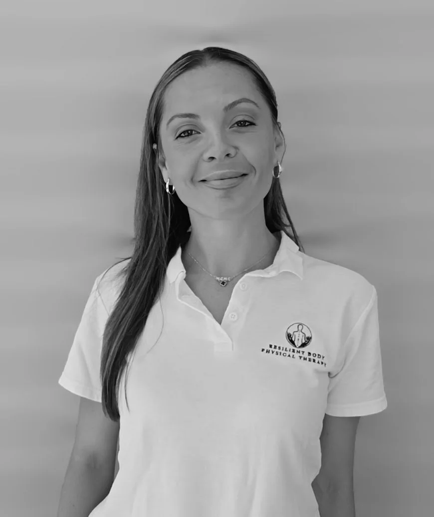 A woman with long hair wearing a white polo shirt with a logo that reads 'Resilient Body Physical Therapy' and earrings, standing against a plain background, smiling gently.