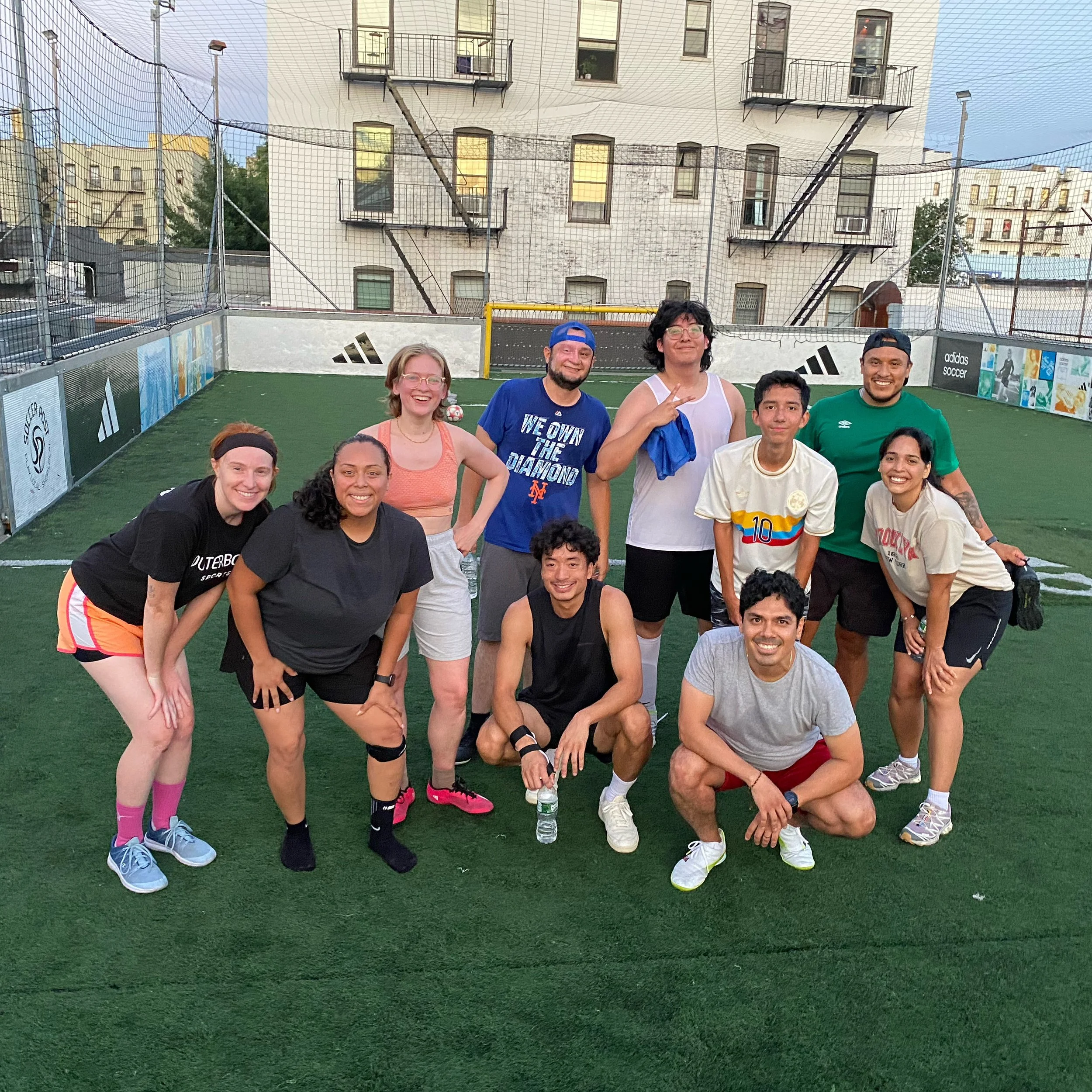 A group of ten people posing together on a small outdoor soccer field, smiling and dressed in athletic wear. The background shows a cityscape with city buildings and a net surrounding the field.