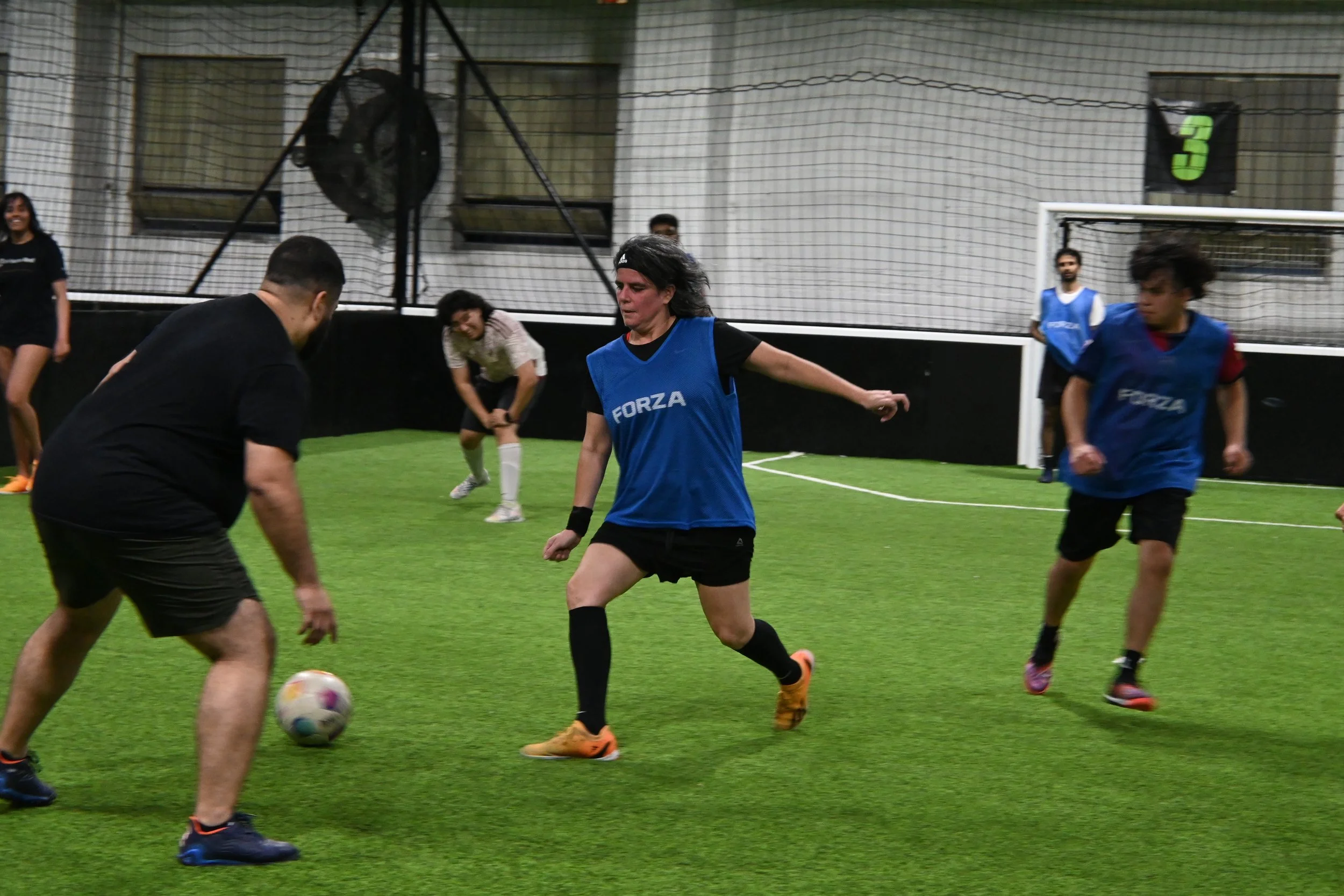People playing indoor soccer practice or game on a green turf field, with some players wearing blue bibs labeled 'FORZA', and a soccer goal in the background.