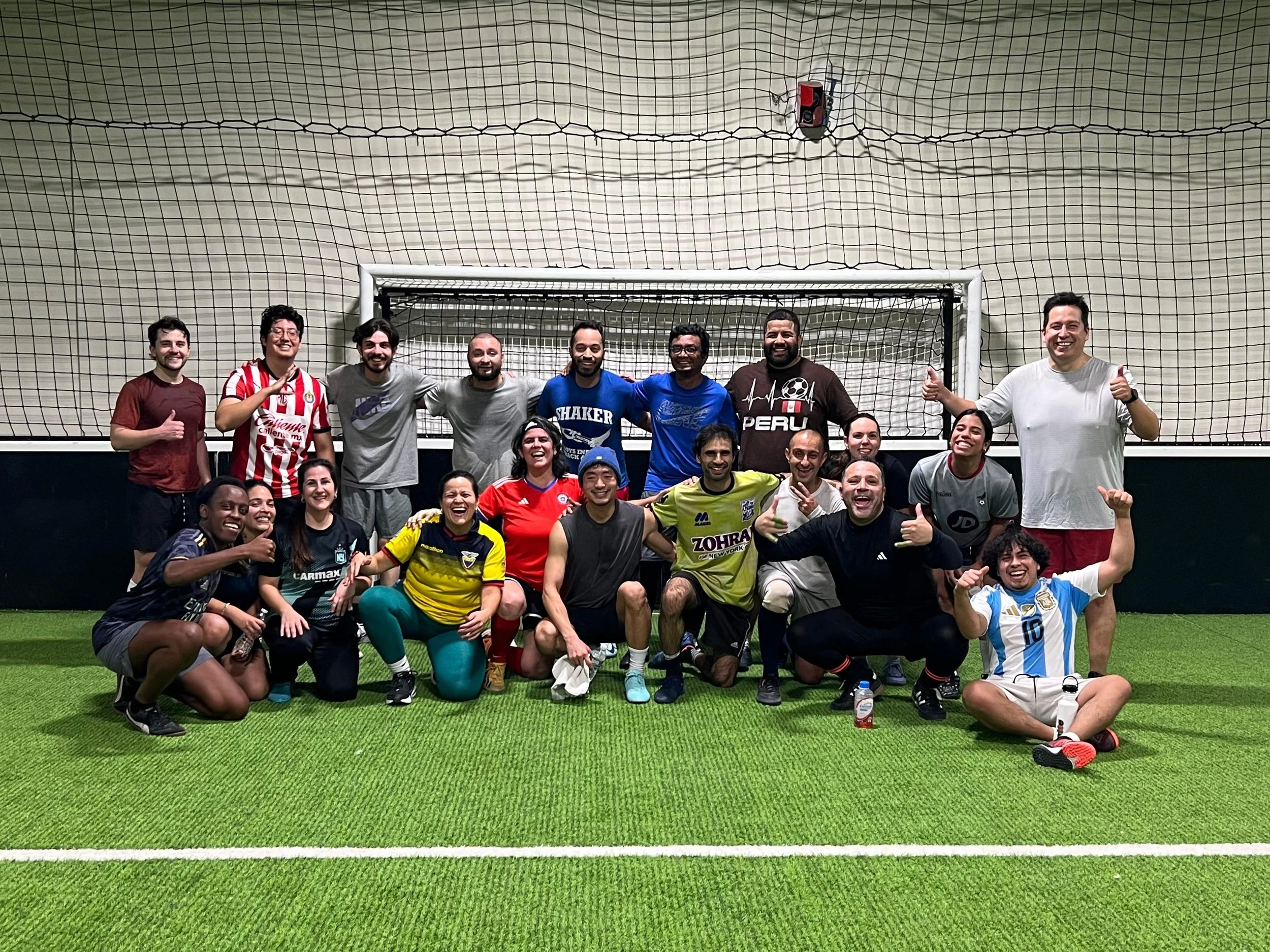 Group of people in soccer jerseys and casual clothes posing on an indoor turf field in front of a soccer goal, celebrating after a game.