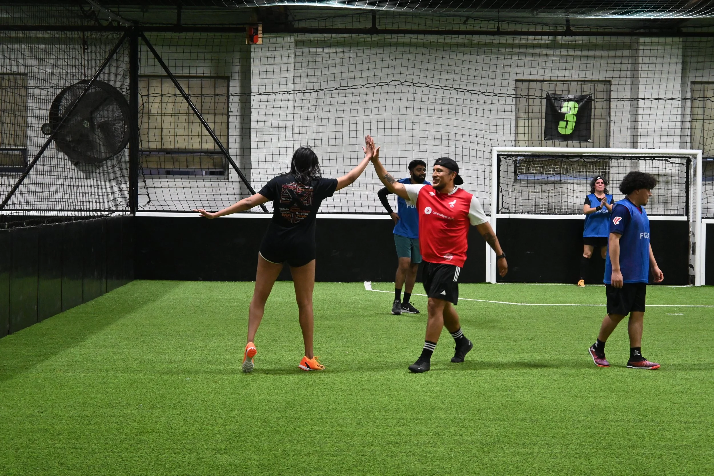 Group of people playing indoor soccer, with some giving high five, in a facility with green artificial turf, netting, and a goal.