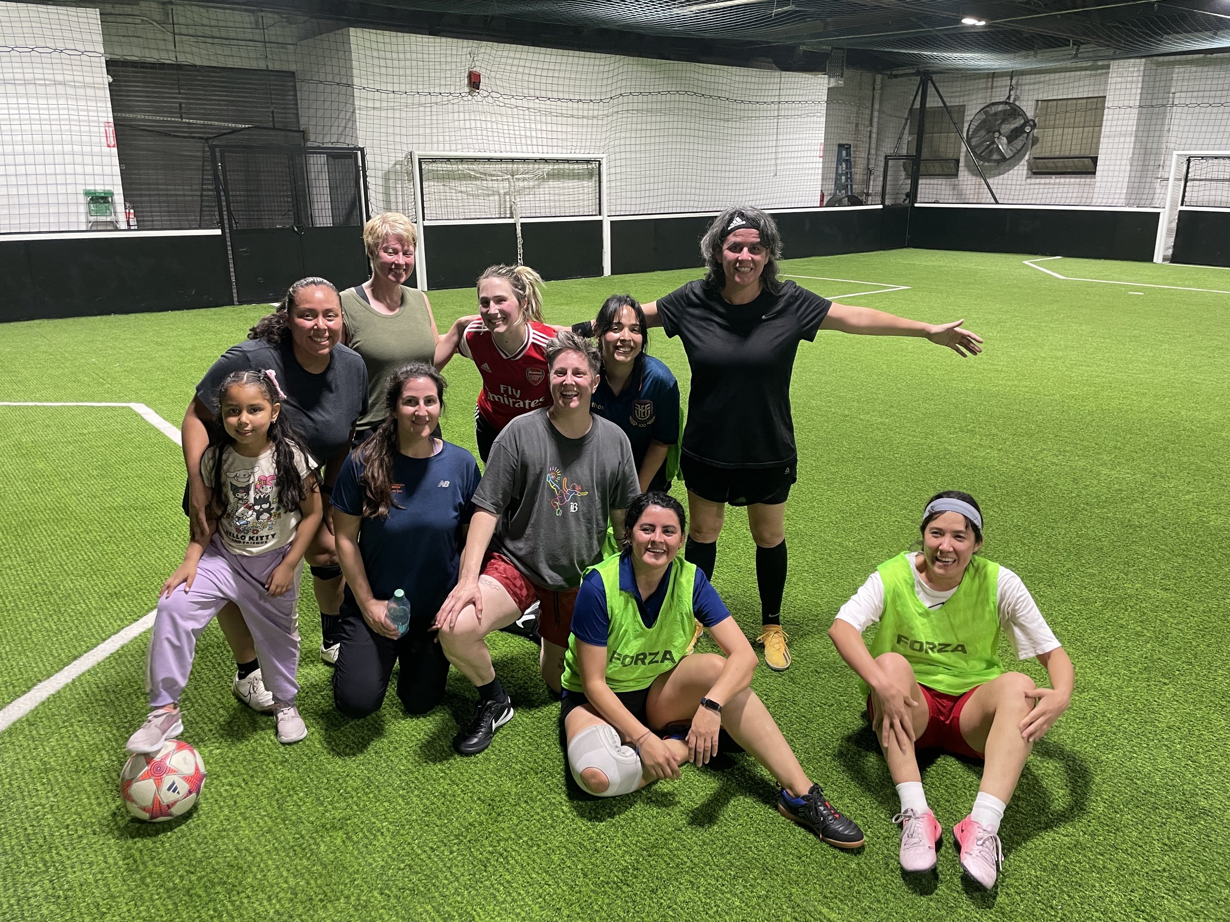 Group of women and girls on an indoor soccer field, smiling for a group photo. Some are wearing athletic gear, with a soccer ball on the ground nearby.