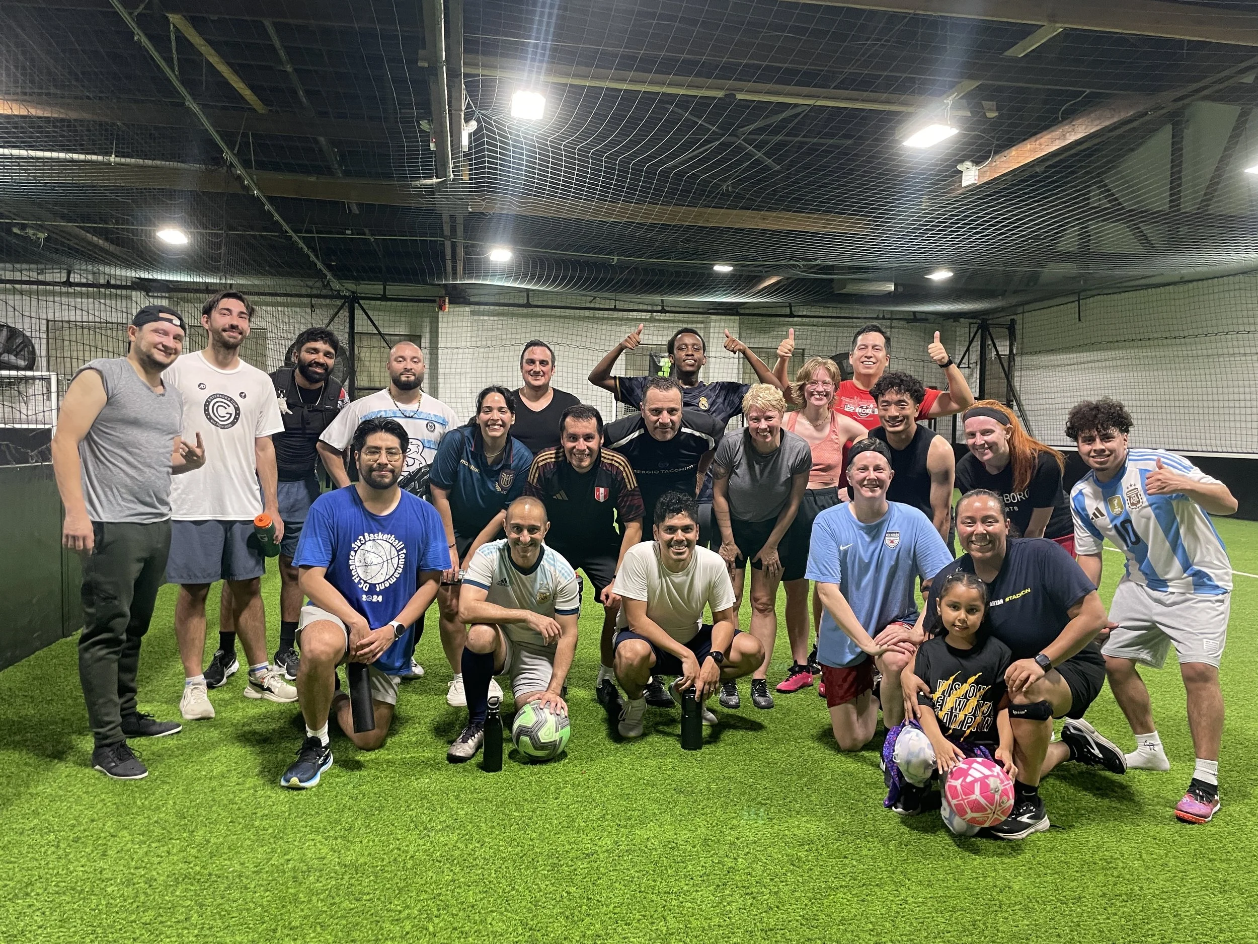 Group of diverse people posing on an indoor soccer field, some giving thumbs up, with soccer balls and water bottles in front, smiling and celebrating.