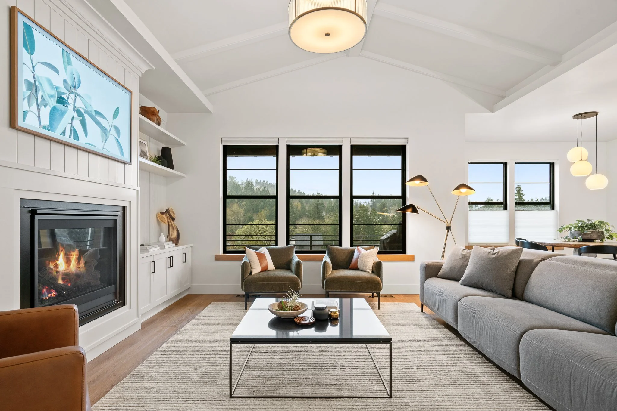 Living room with a gray couch, two green armchairs, a black coffee table, a fireplace, and large windows showing outdoor greenery
