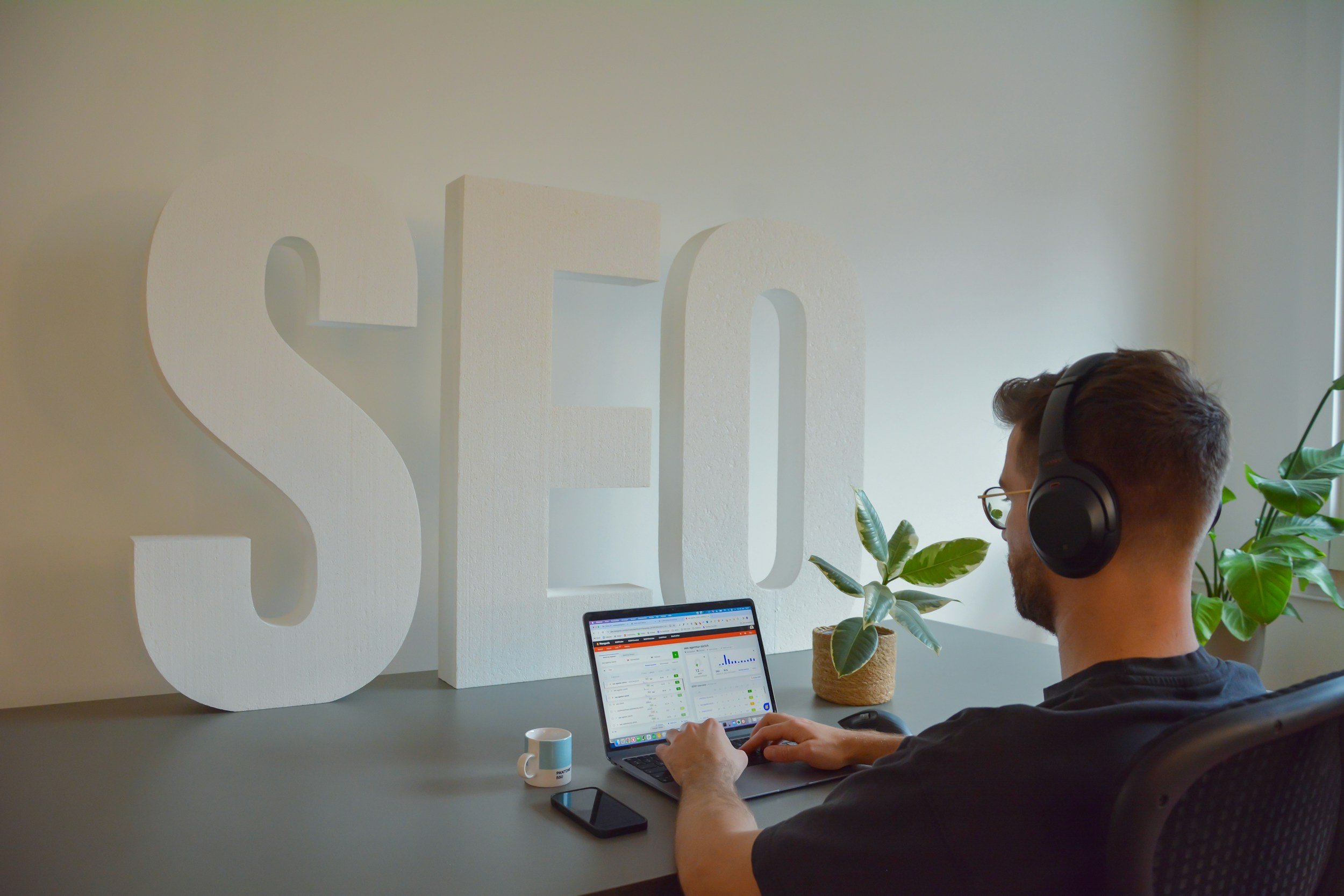 A man wearing glasses and headphones working on a laptop at a desk with plants, a coffee mug, and a smartphone in front of large white letters spelling 'SEO' on the wall behind him.