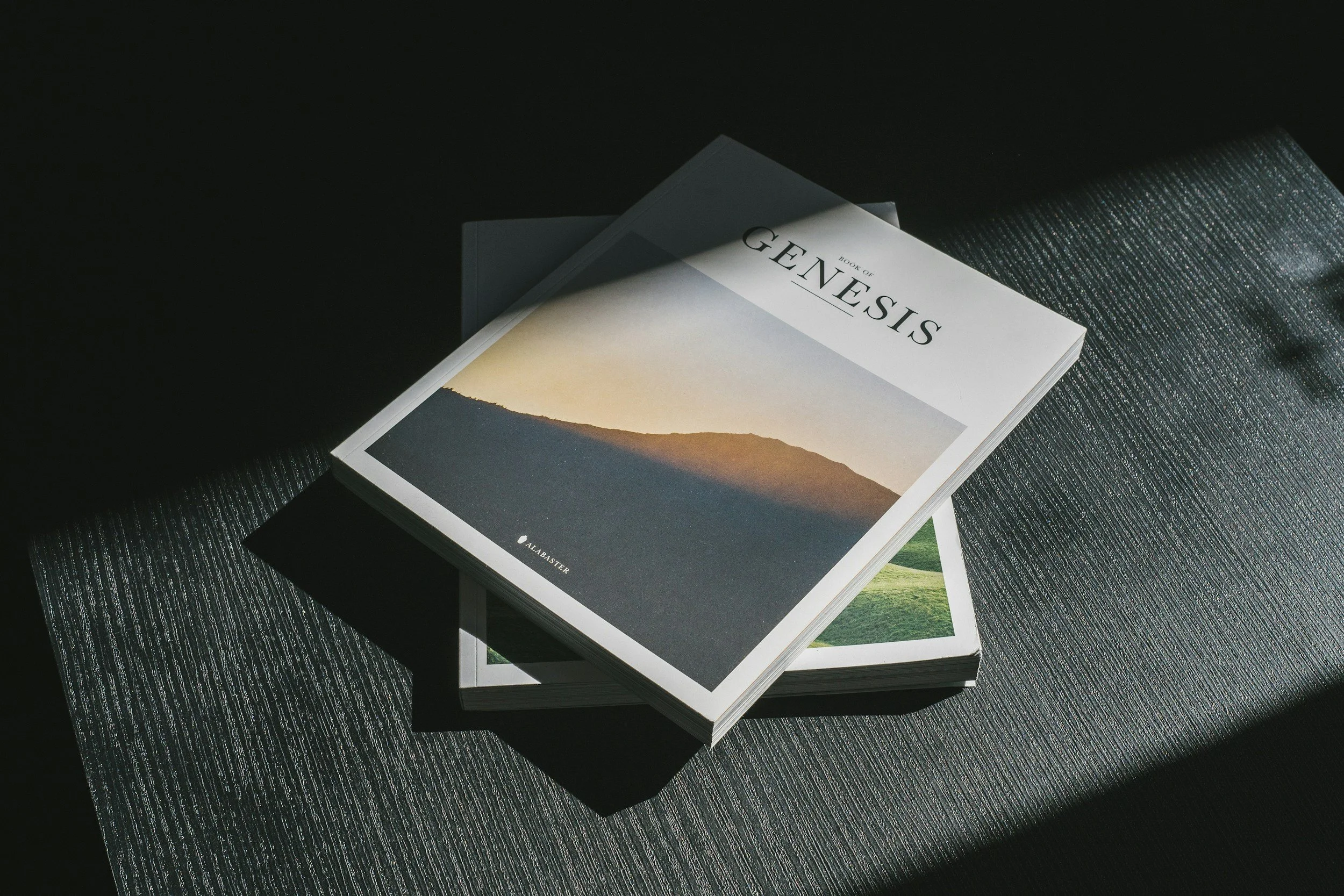 A book titled 'Genesis' resting on another book, placed on a textured dark surface with lighting creating shadows.