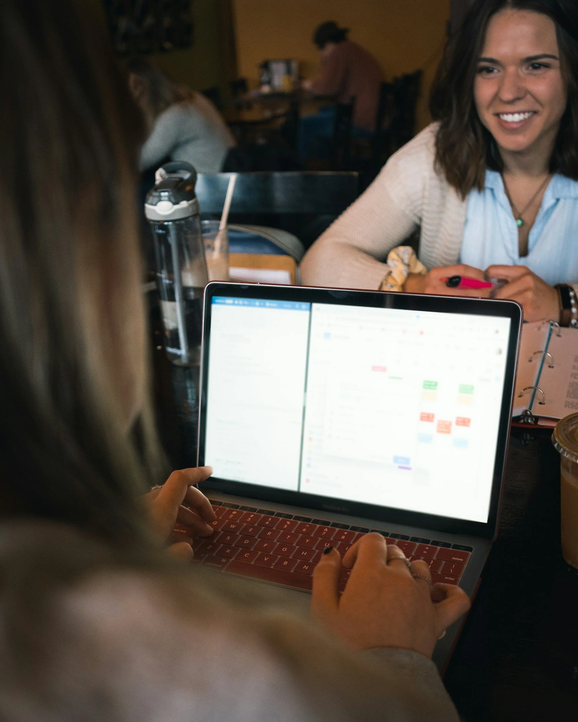 Two women sit at a table in a cafe, one is using a laptop with a red keyboard and the screen showing a calendar or planner, while the other woman is smiling and holding a pink pen. There is a glass of iced coffee and a water bottle on the table.