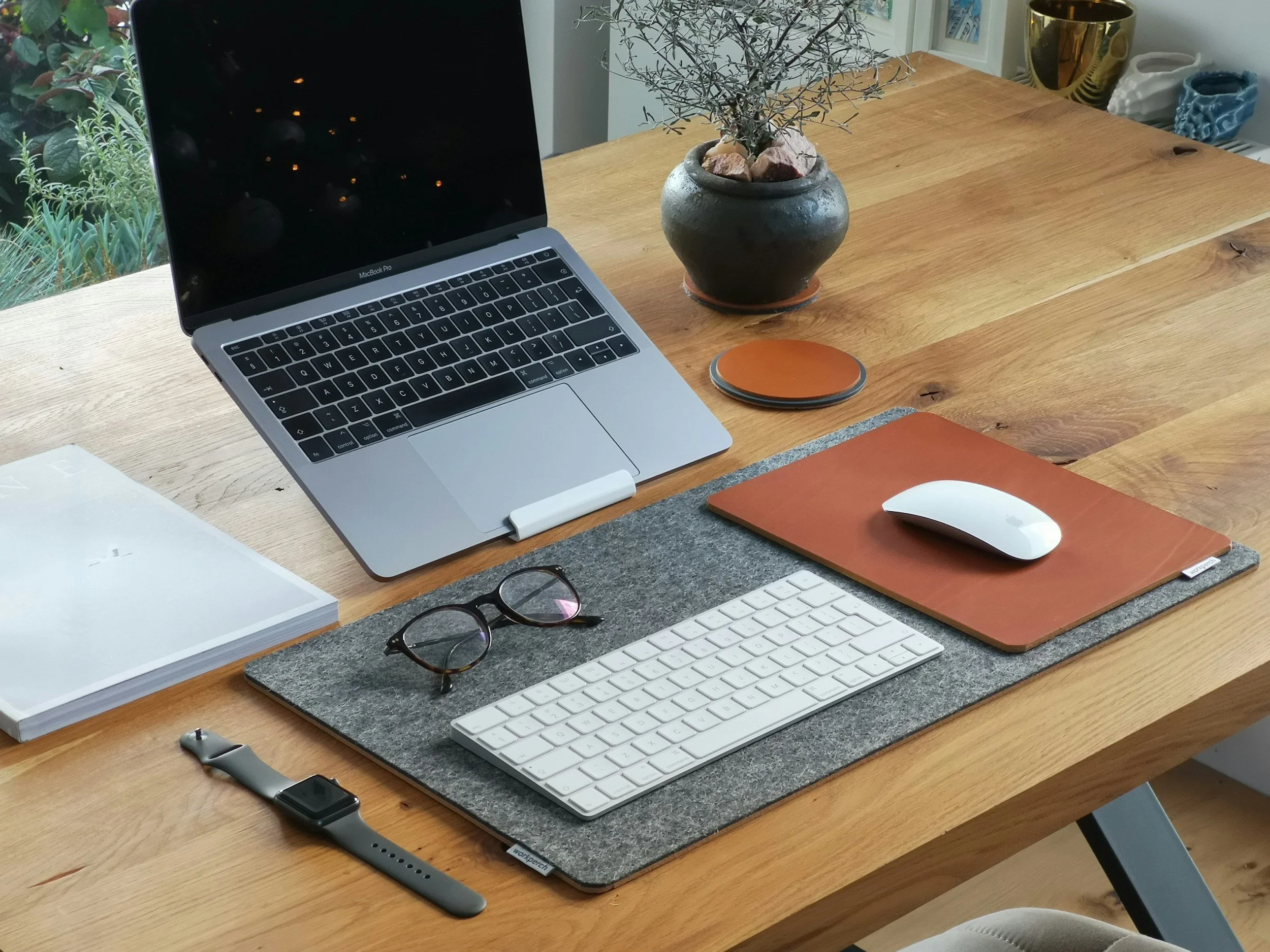 A tidy wooden desk with a MacBook Pro laptop, a wireless keyboard, a wireless mouse on a brown leather mouse pad, a pair of glasses on a gray felt desk mat, an Apple Watch, a notebook, and a round wireless charger. In the background, there is a potted plant near a window and some decorative objects on shelves.