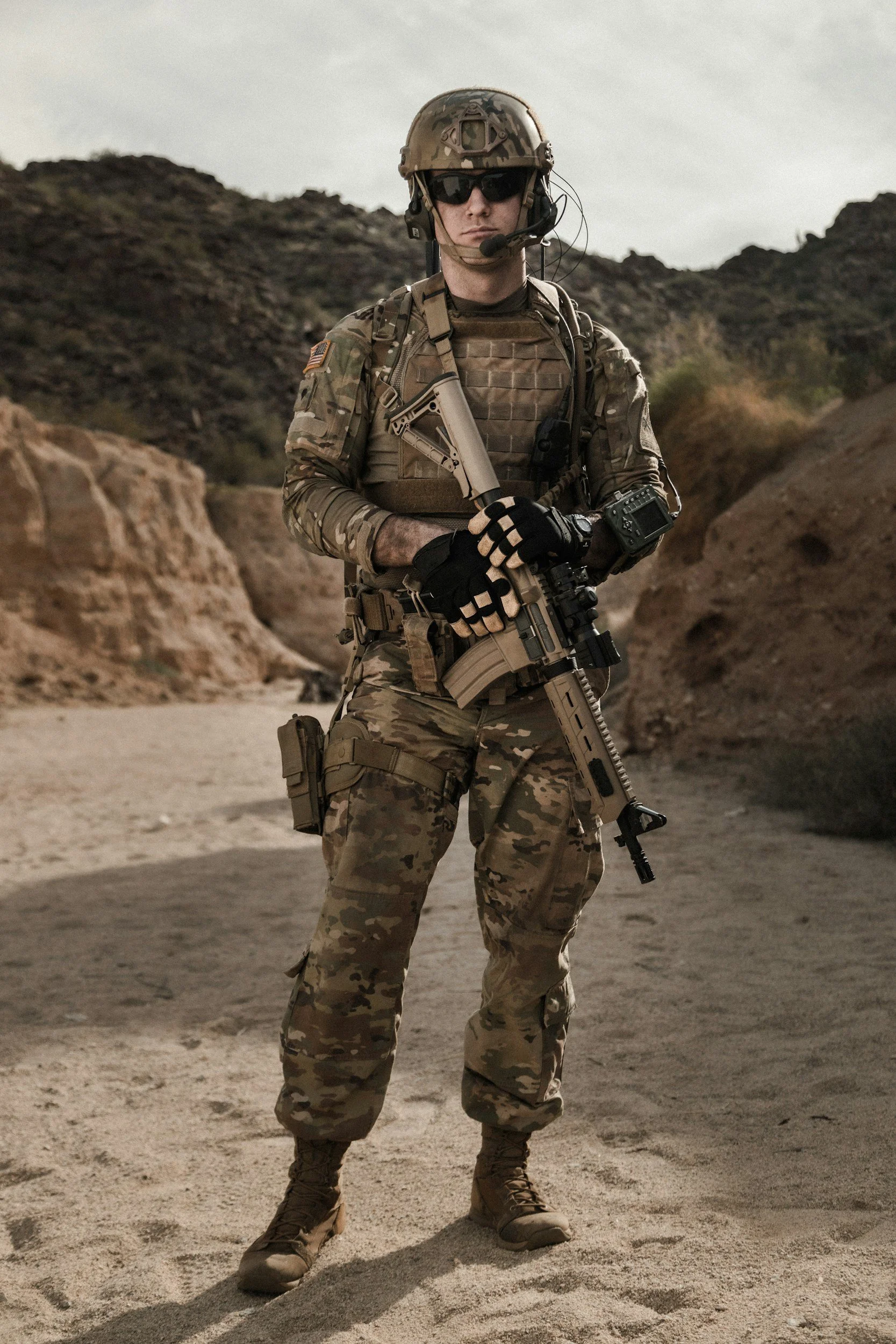 A soldier wearing camouflage uniform, helmet, sunglasses, and tactical gear standing in a desert landscape with rocky hills.