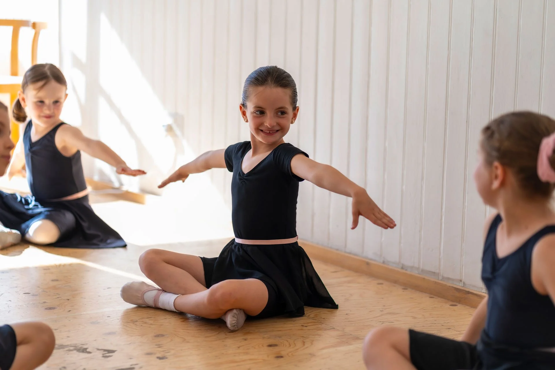 Young girls practicing ballet in a studio with wooden floors and white paneled walls, wearing black leotards and tights, sitting on the floor, with one girl in the center smiling and extending her arms.
