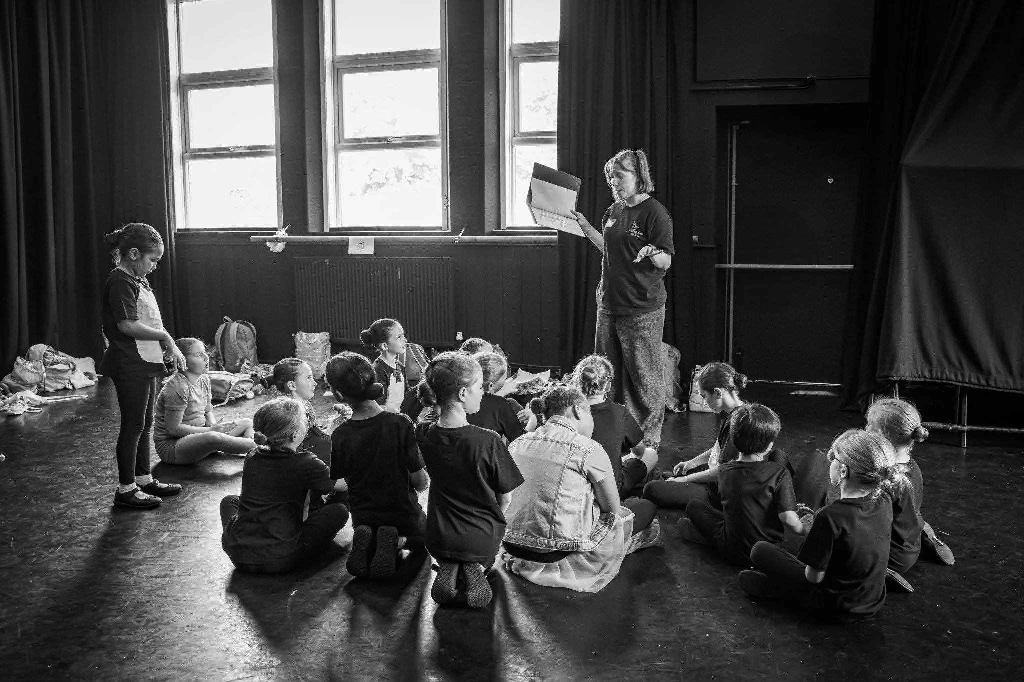 A group of young girls sitting on the floor listening to an instructor in a room with large windows and black curtains.