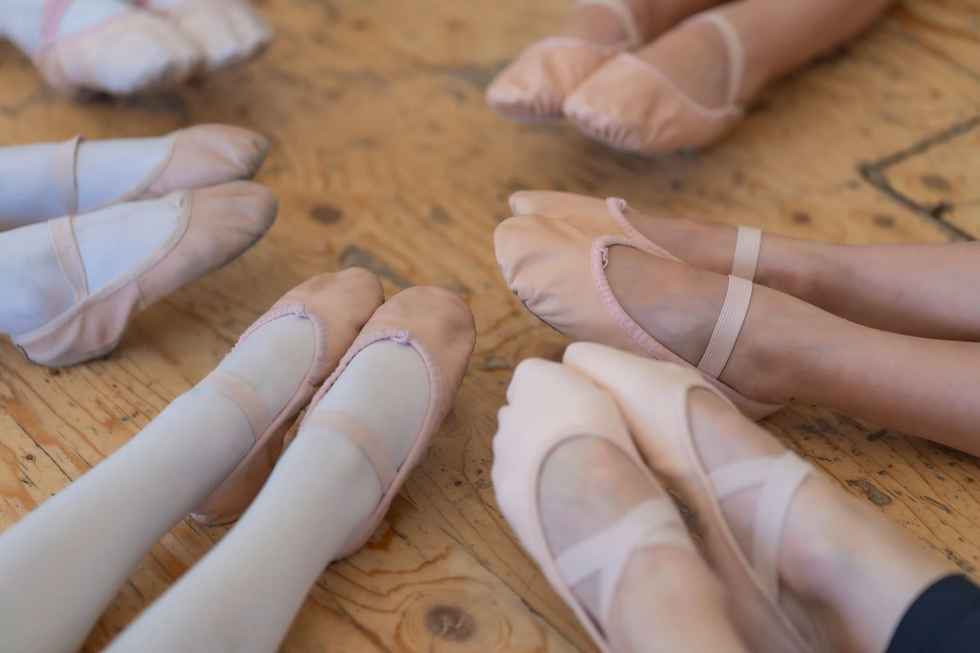 Ballet dancers wearing pink and white pointe shoes sitting in a circle on a wooden floor.