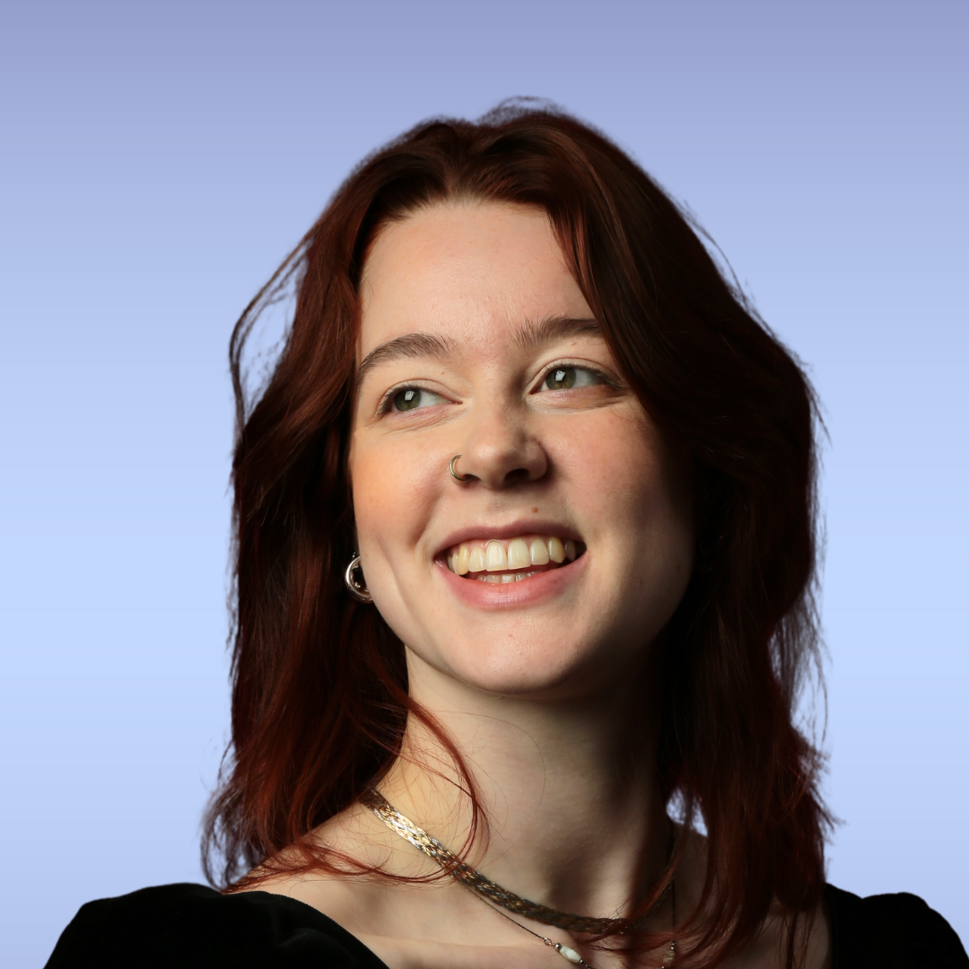 Close-up of a young woman with red hair, blue eyes, and a bright smile, wearing turquoise jewelry and a light blue top against a gradient blue background.