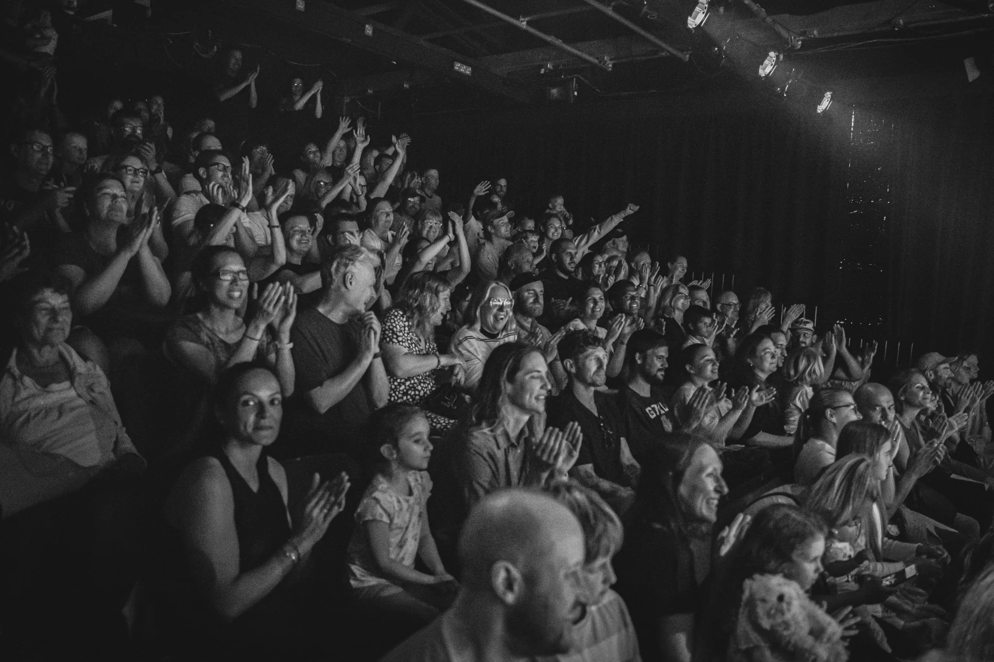 Black and white photo of an audience on a theater or comedy club, with people clapping and smiling, some with hands raised, 다양한 연령대와 인종이 포함되어 있음.