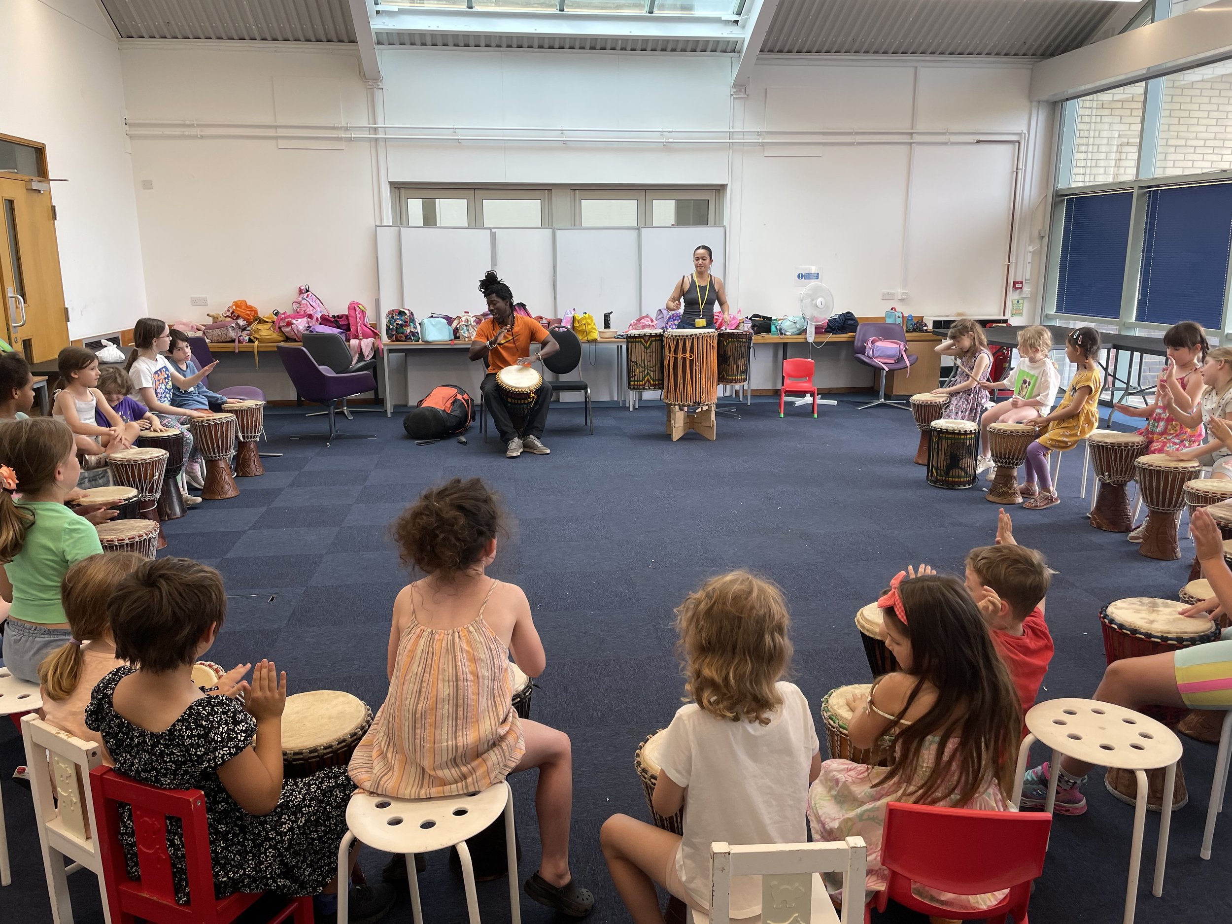 Children sitting in a circle playing drums in a classroom, with two adults leading the musical activity at the front.