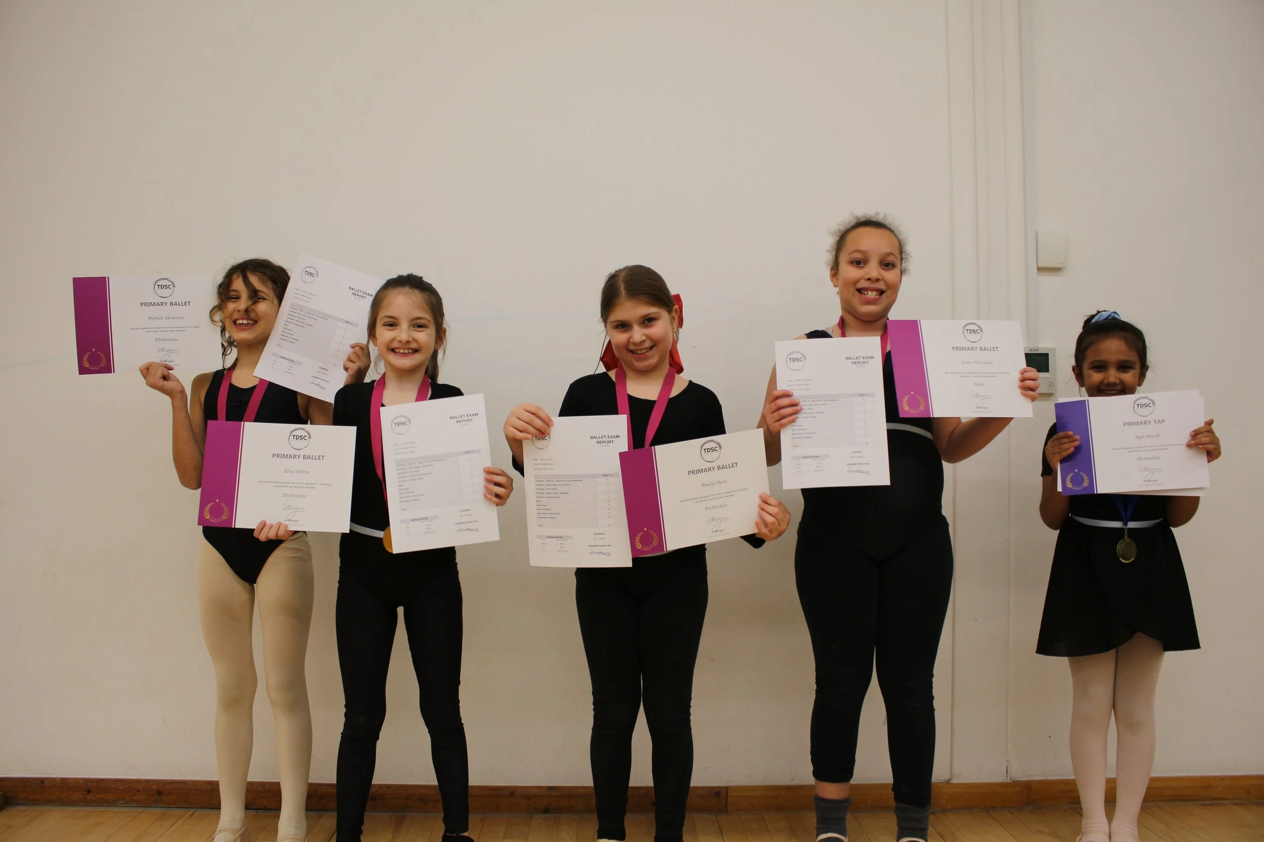 Five girls in smart dance uniforms smile and look proud while holding dance exam certificates and medals.
