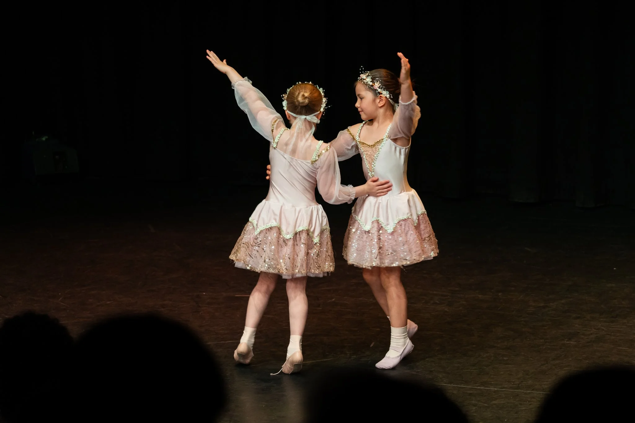 Two young girls in ballet costumes dancing on stage, holding each other with arms raised, dark background, stage lighting.