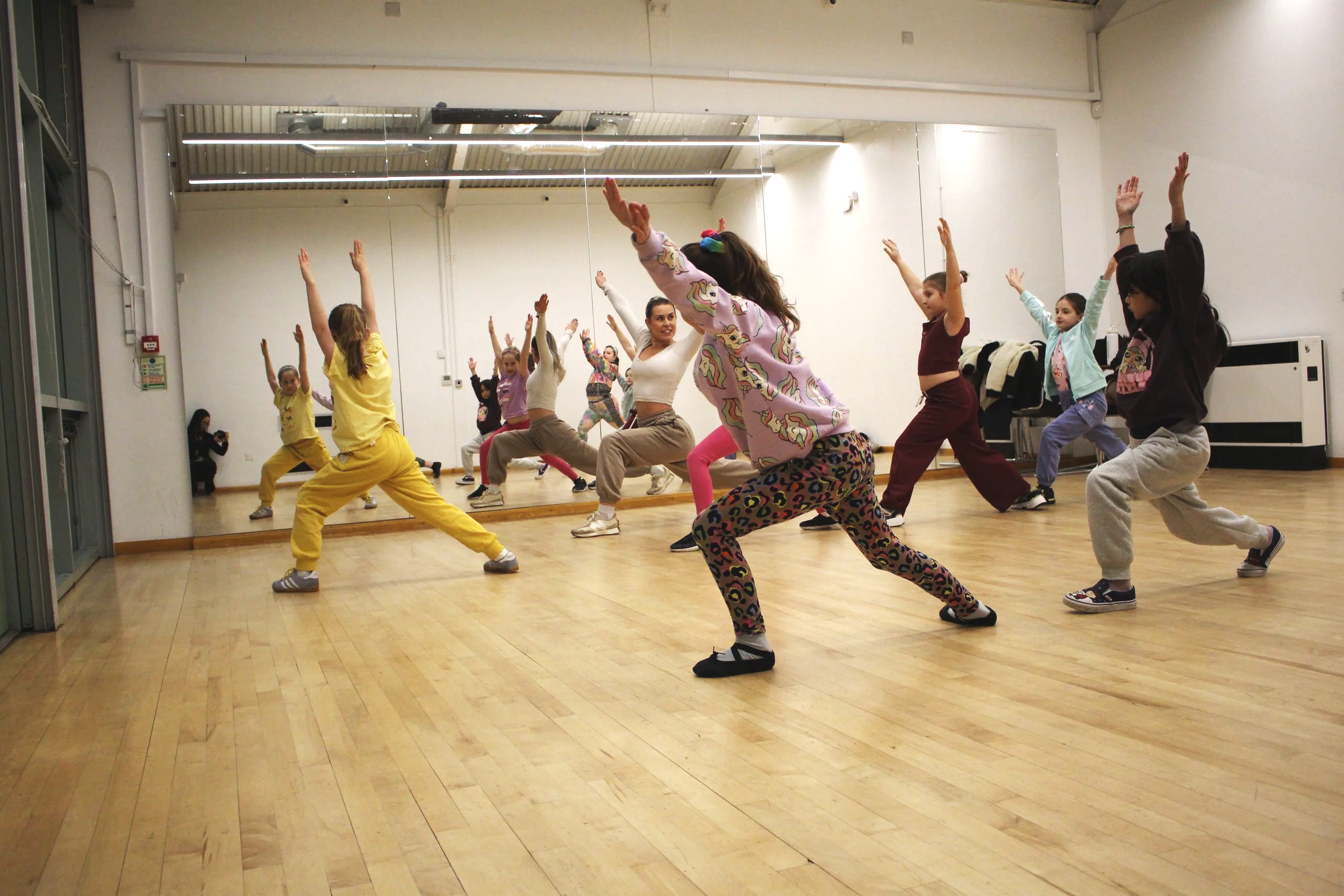 A group of kids reach their arms into the air while in a lunge. Their teacher smiles and leads the class from the front of the room.