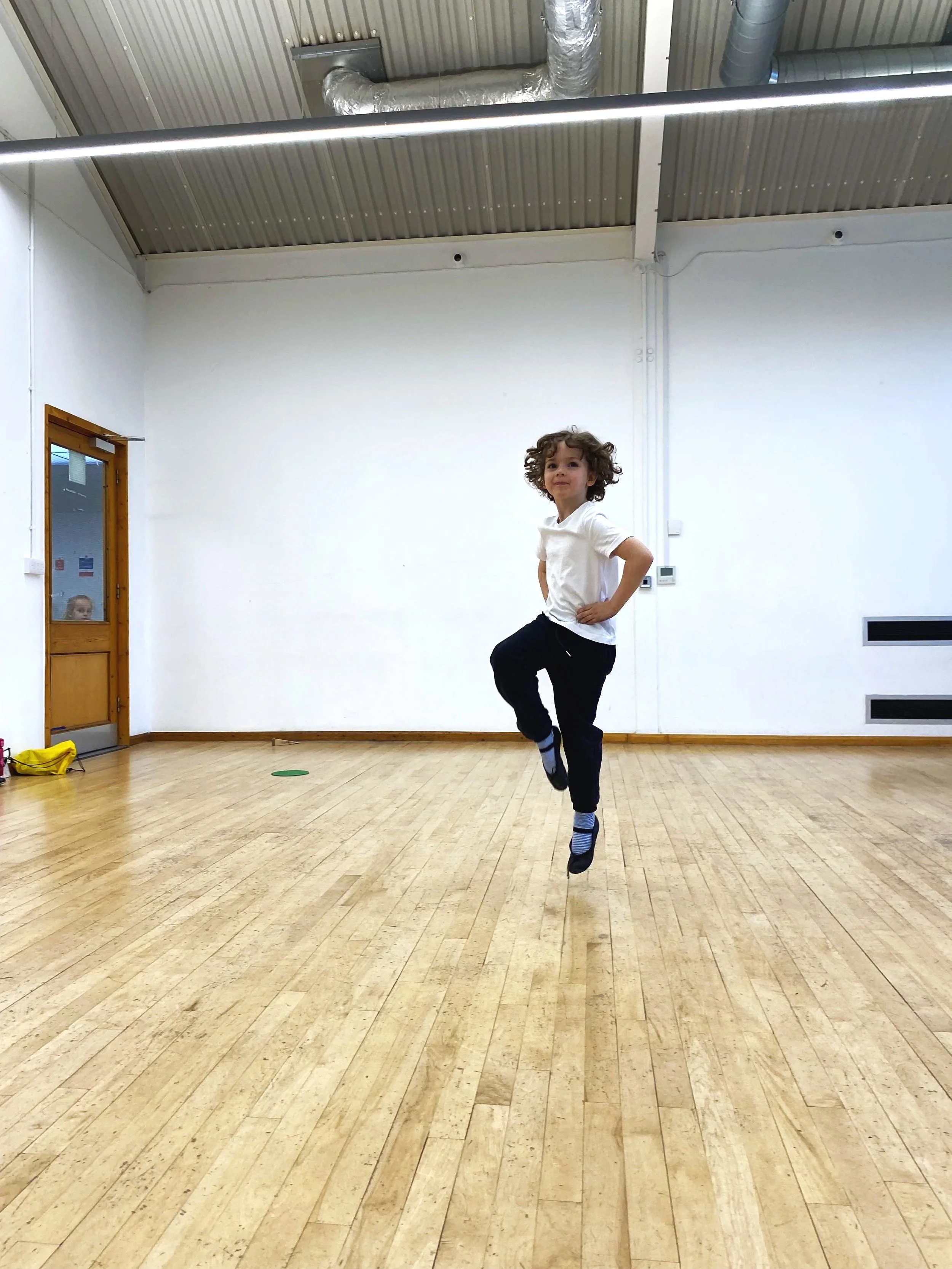 A happy young boy keeps into the air on one leg with his hands on his hips.