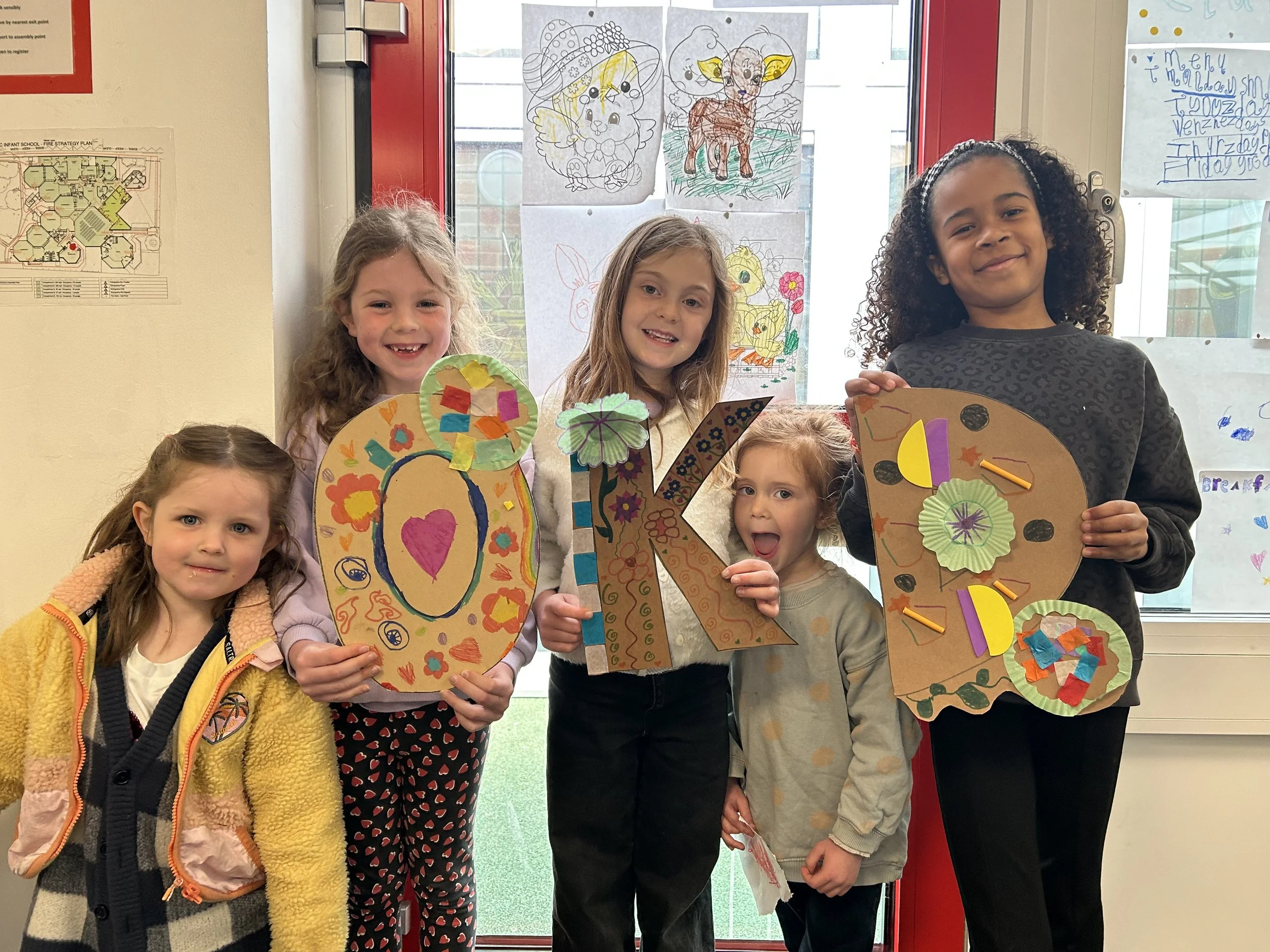 Six children standing in a row indoors, holding decorated letter cutouts spelling 'I K D'. They are smiling and appear to be in a classroom or art room, behind them are colored drawings of animals on the window. Each child is wearing casual clothing, and they seem to be proud of their artwork.