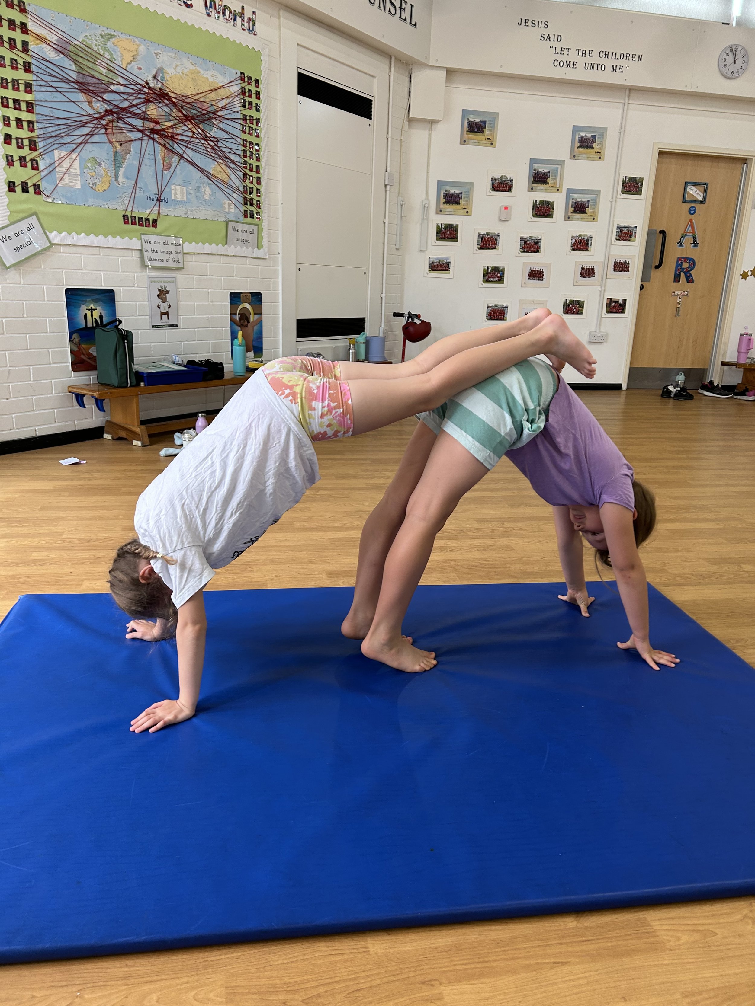 Two children performing a partnered yoga pose on a blue mat in a classroom, with one child bending back with hands and feet on the ground, and the other child supporting them by holding their hips.