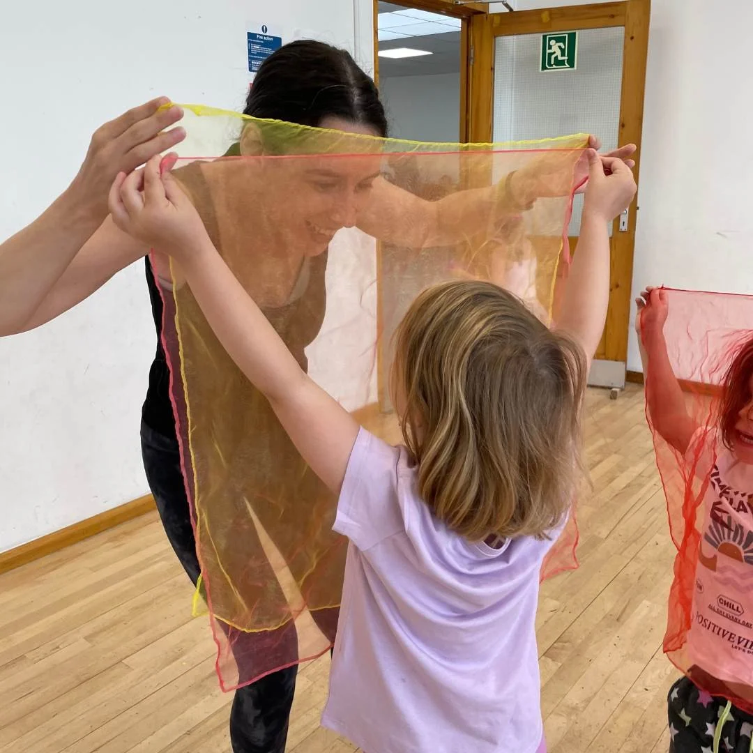 A woman and two children holding colorful sheer scarves in a room with wooden floors and walls, engaging in a playful activity.