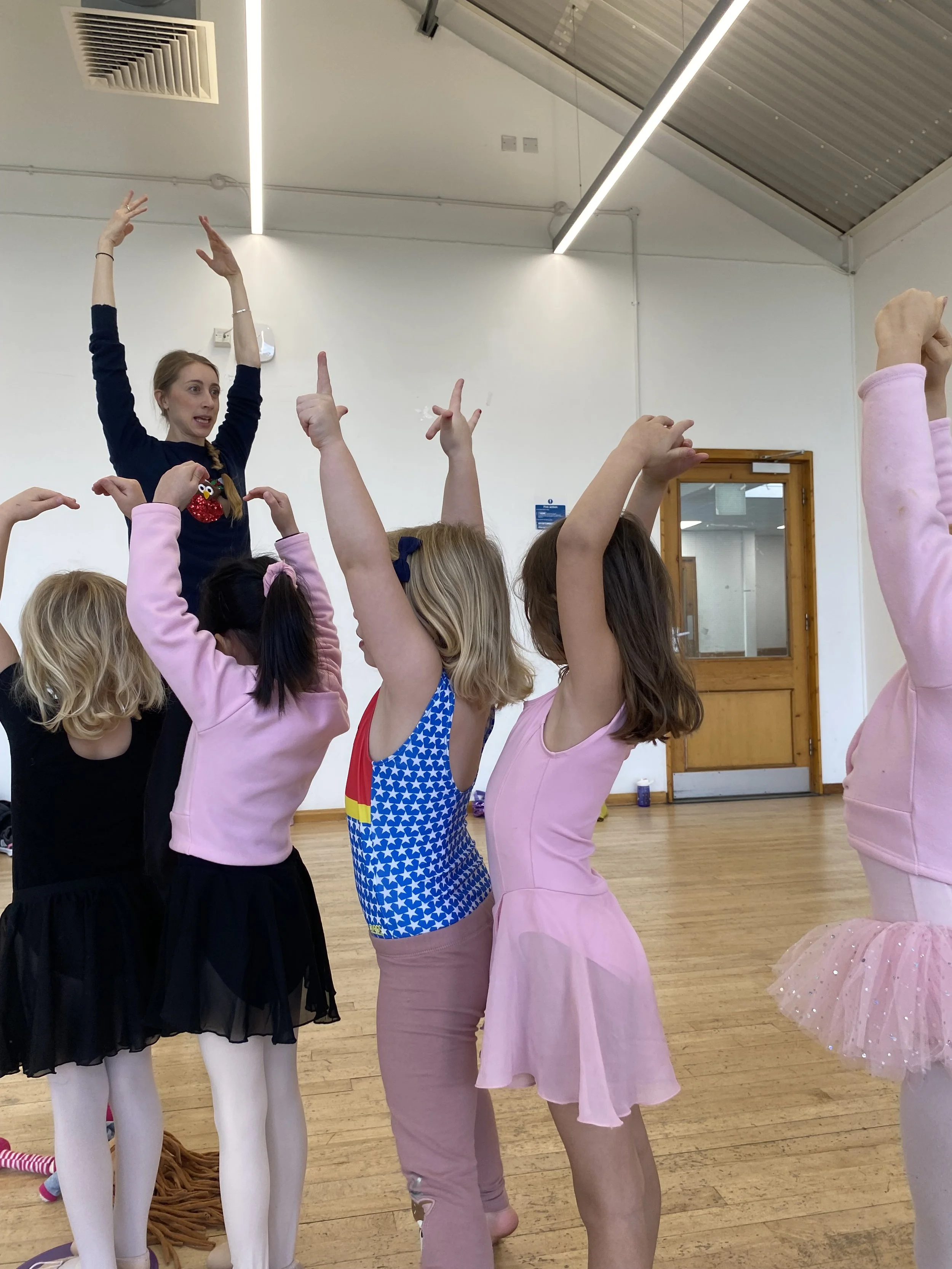 A ballet teacher leads of group of young girls in ballet outfits in an arm exercise.