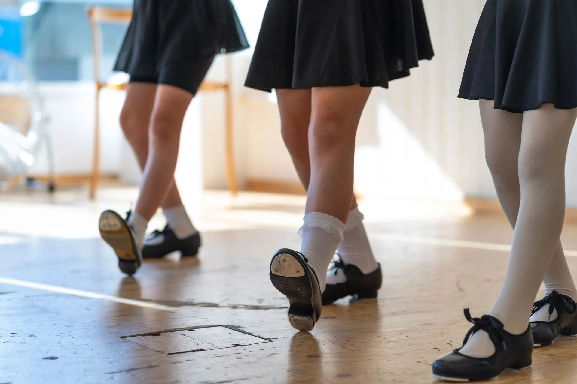 Three people dancing in black skirts and white socks with black shoes, captured from behind.