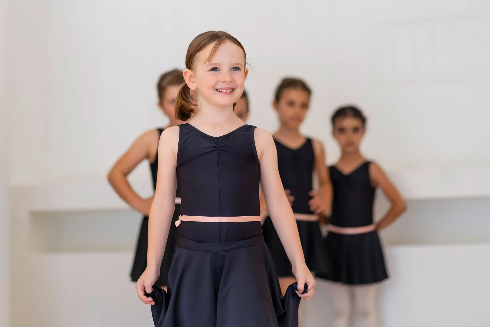A young girl in a navy ballet costume smiling, standing in front of four other girls in similar costumes with their hands on their hips in a dance studio.