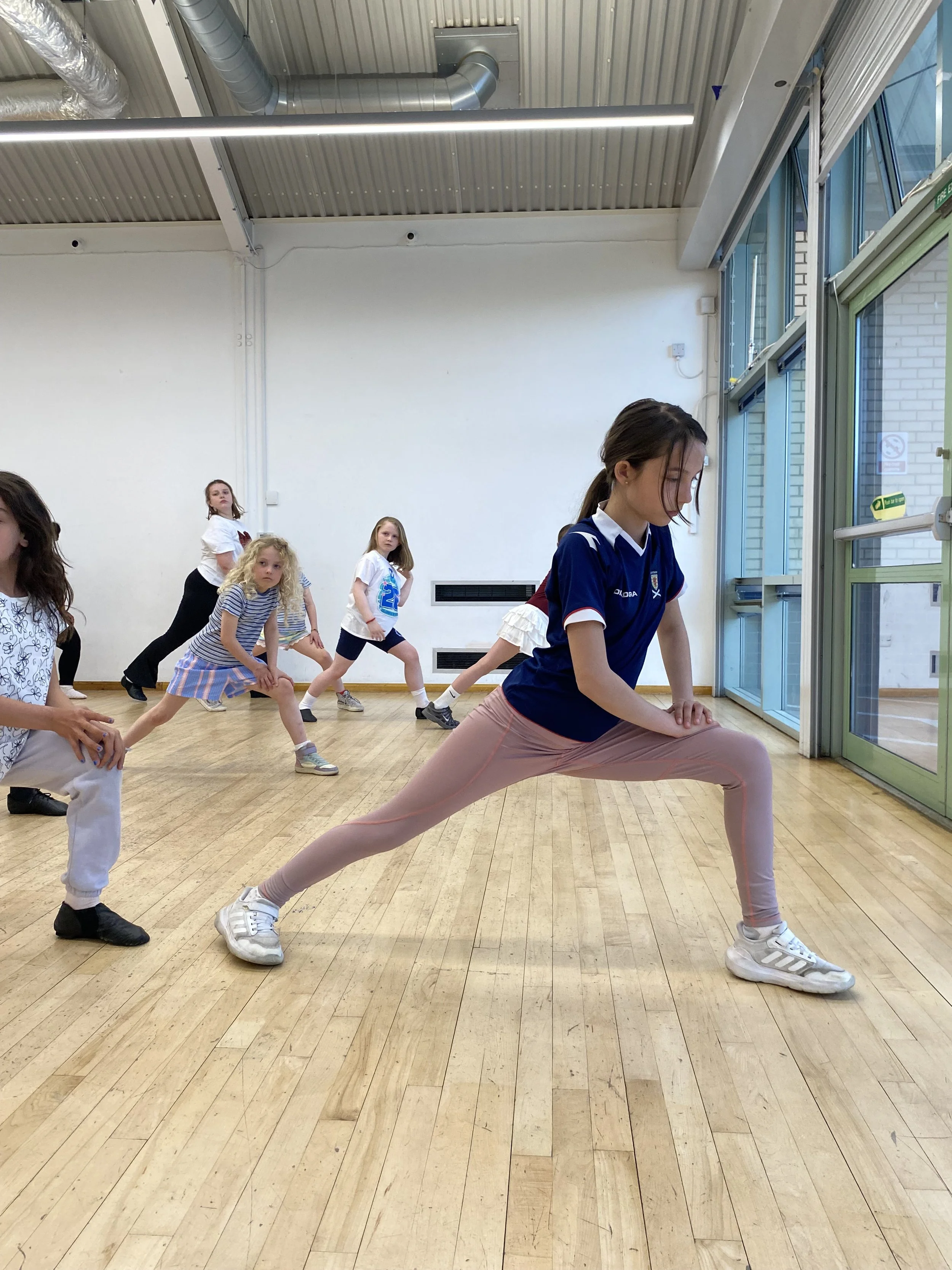 A girl stretching in a dance studio doing a split with one leg extended in front and the other behind, on a wooden floor, with a mirror reflecting her and other children in the background.