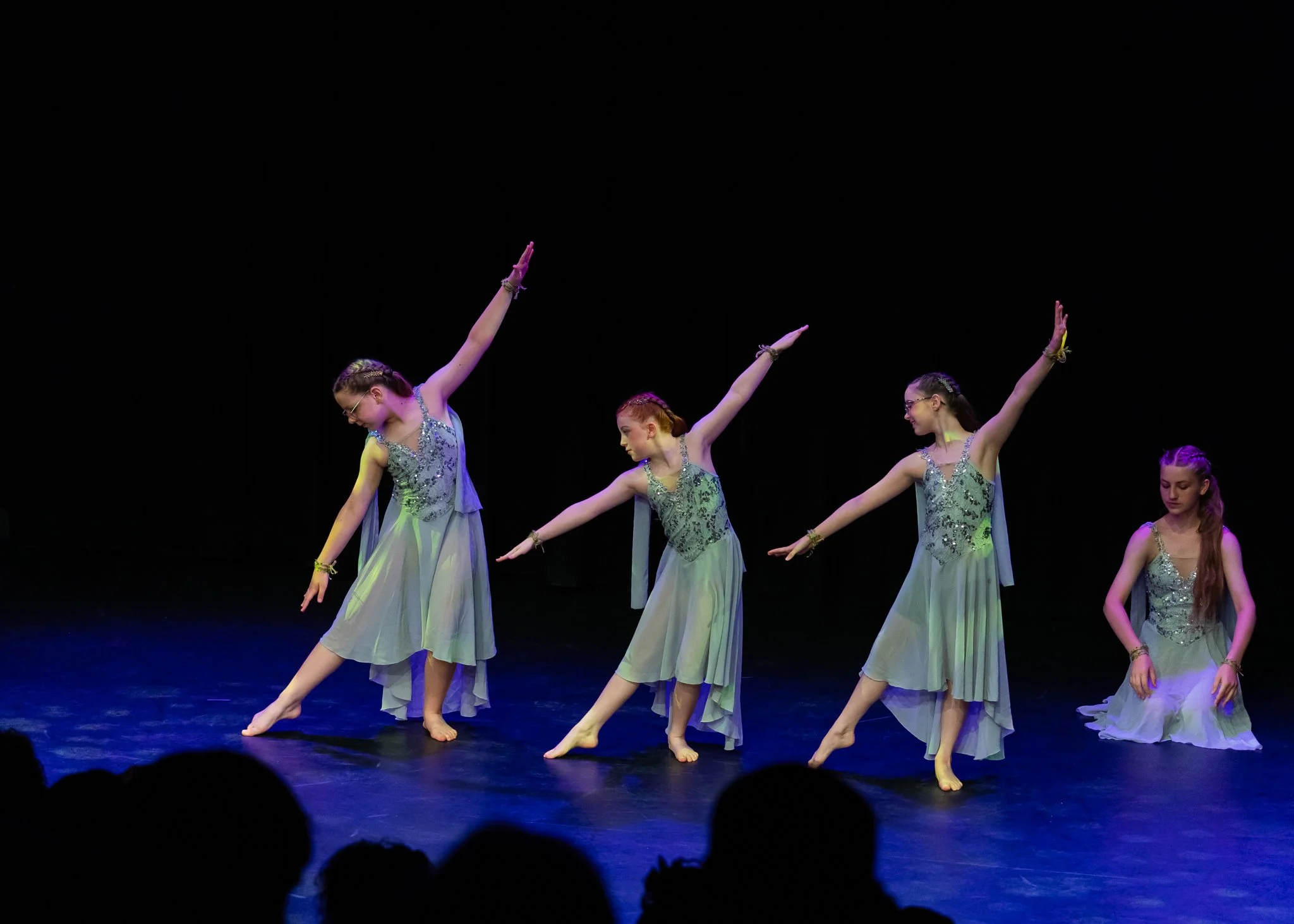 Four young girls in silver and lavender dance costumes performing a ballet on stage with black background.
