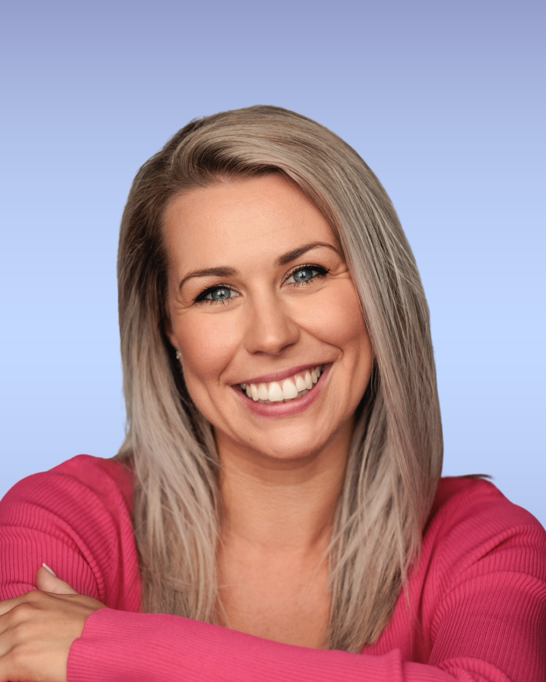 A smiling woman with blonde hair and blue eyes wearing a pink top, posing with her arms crossed against a light blue background.