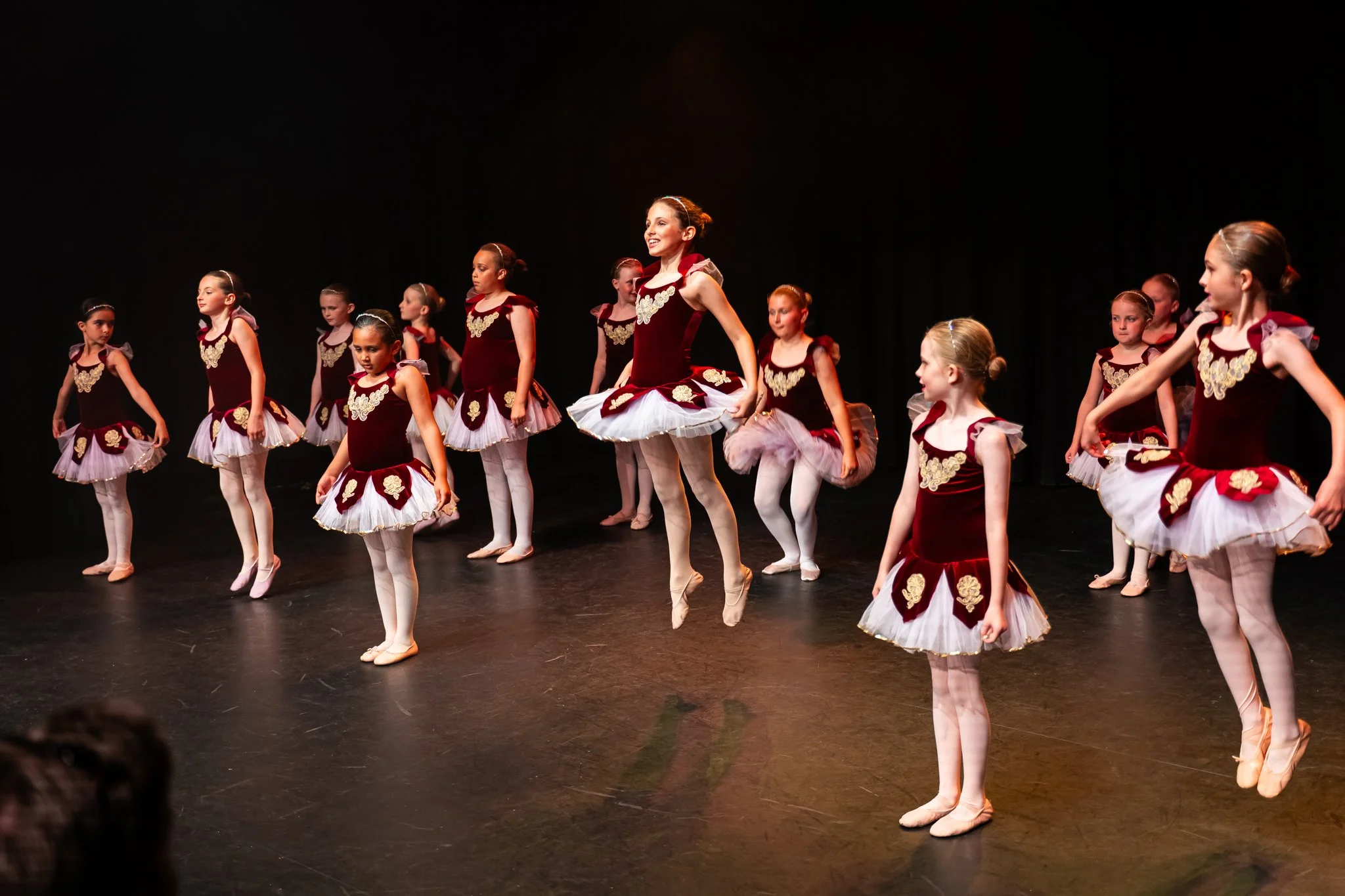 Group of young ballet dancers on stage, some jumping and others standing, dressed in maroon and white costumes with gold embellishments, performing a dance.
