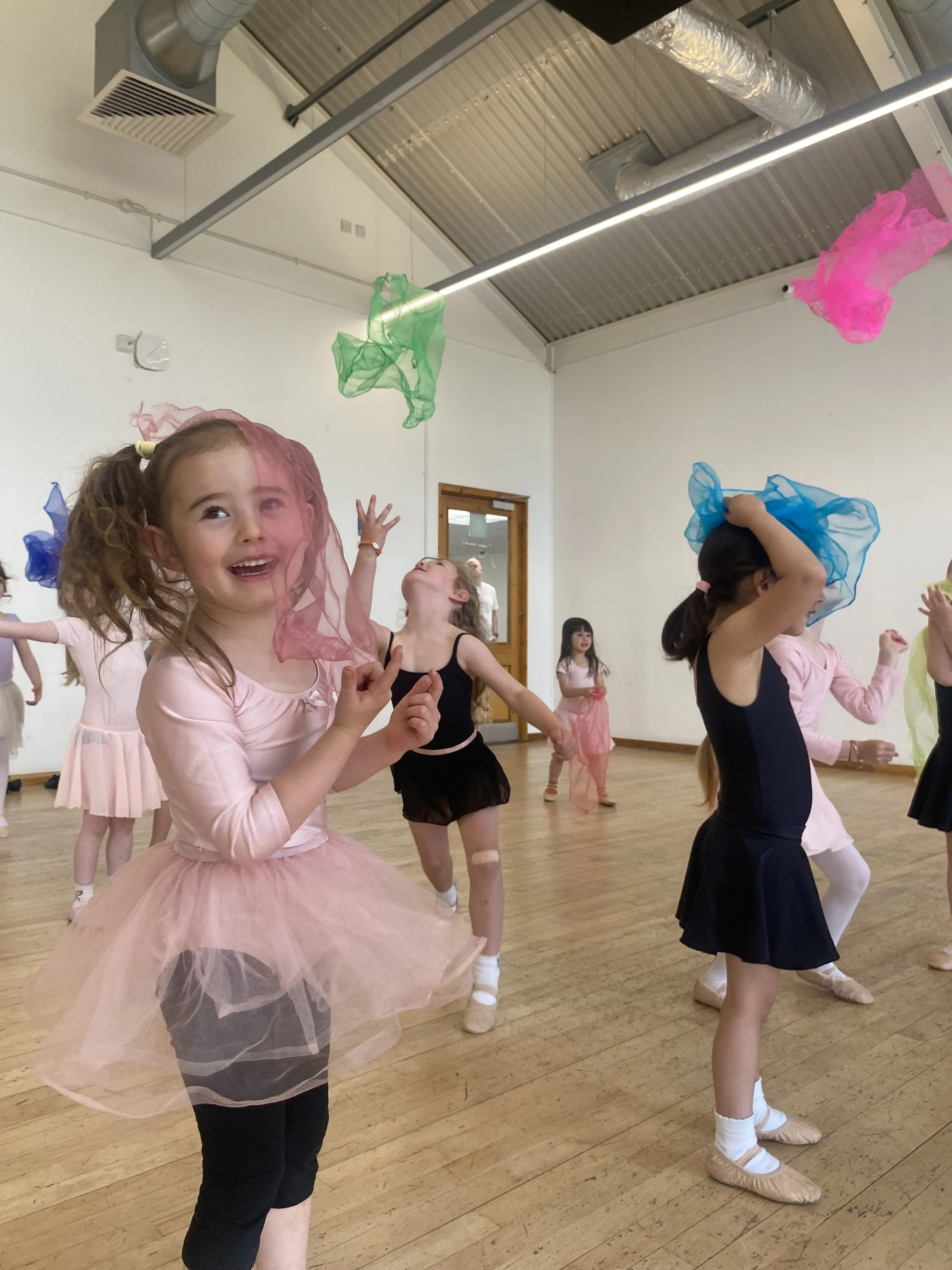 A young girl in a black dress, pink cardigan, and gray tights is standing inside a dance studio on a yellow disc, with several other colored discs scattered on the wooden floor. In the background, a group of other young girls are standing near a mirror, wearing pink dresses, and some are practicing dance moves.