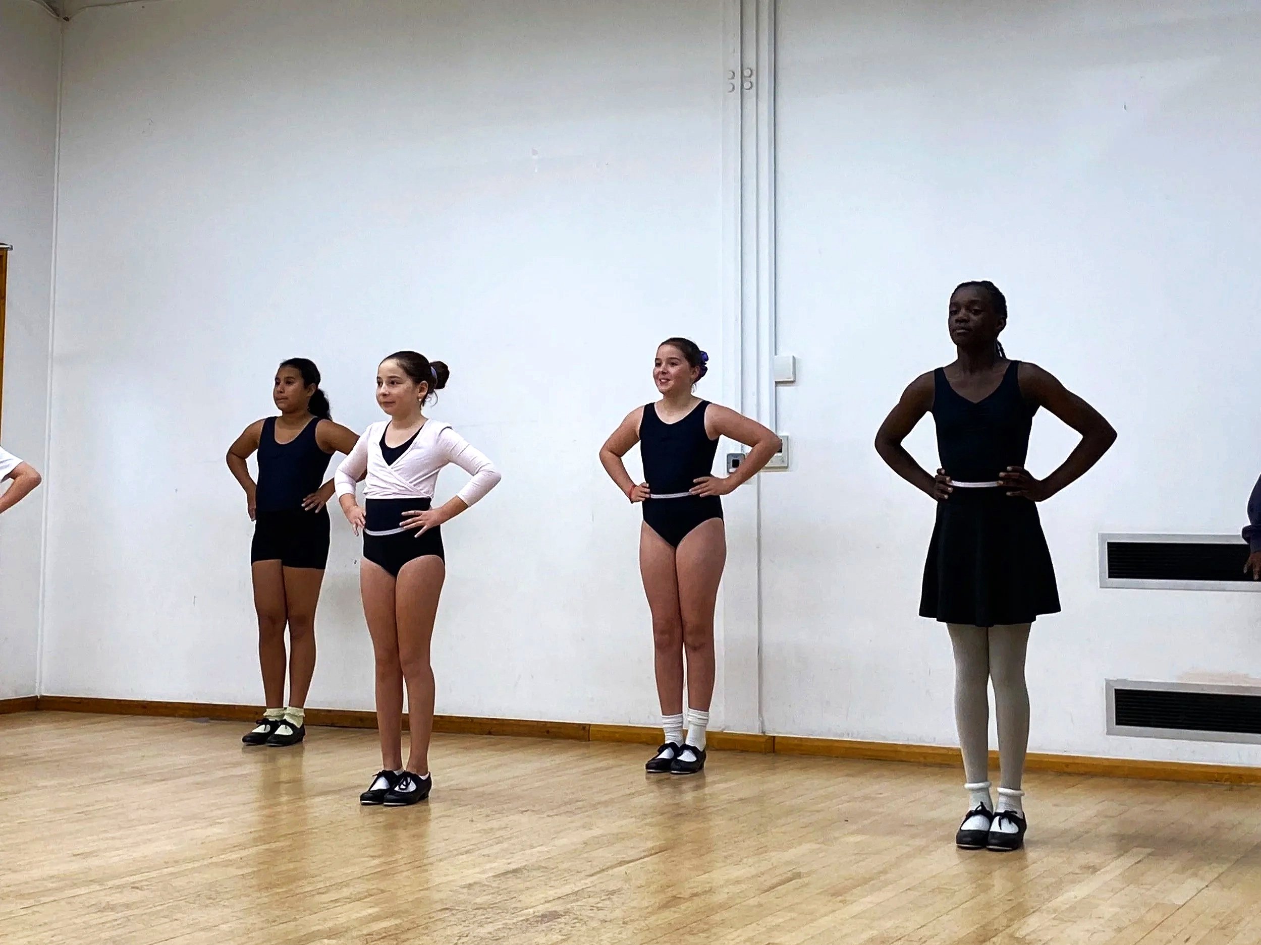 Four young girls practicing ballet in a dance studio, standing with hands on hips, wearing dance costumes and slippers.