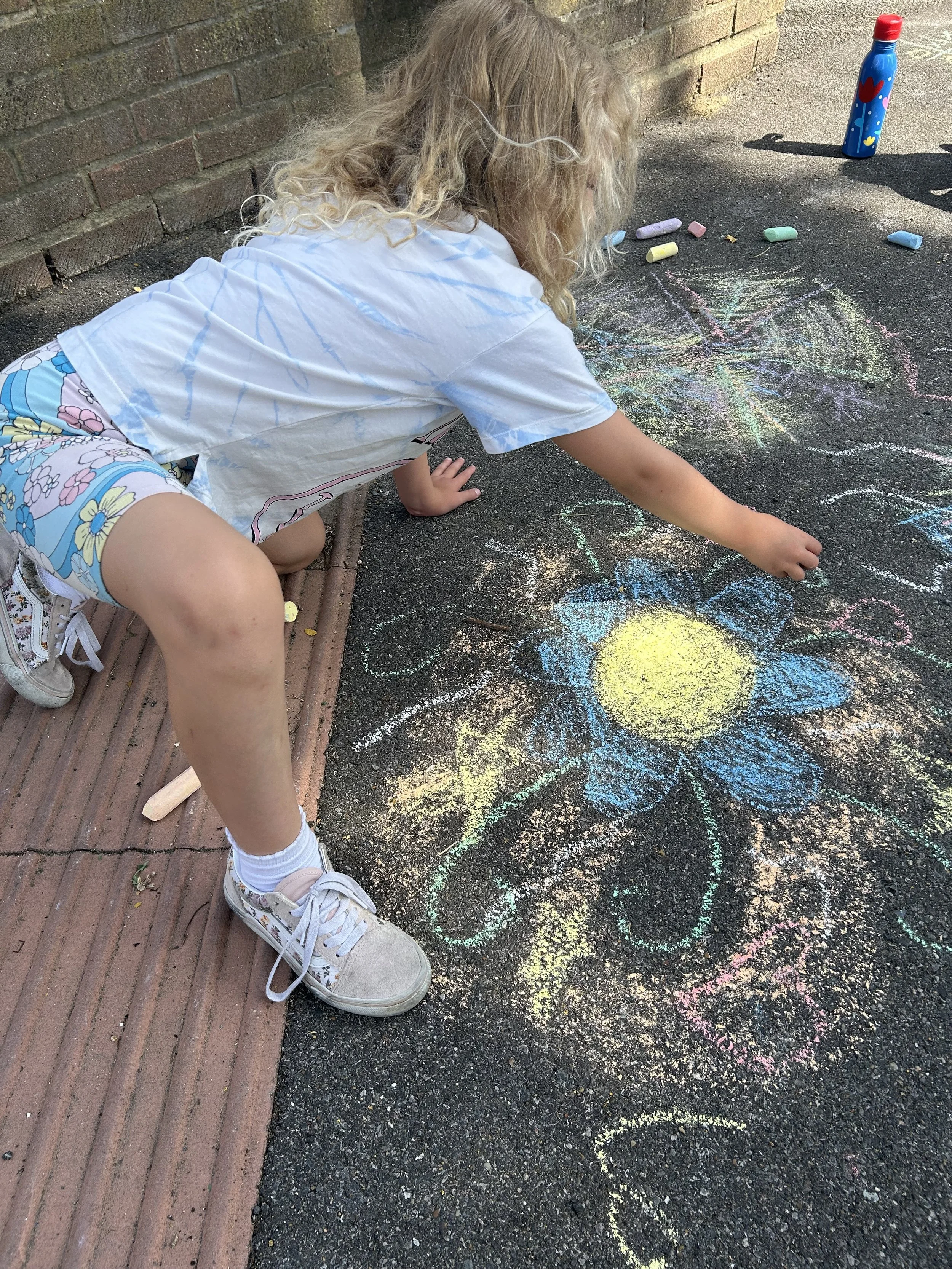 A young girl with curly blonde hair drawing a large yellow and blue flower with chalk on a sidewalk.