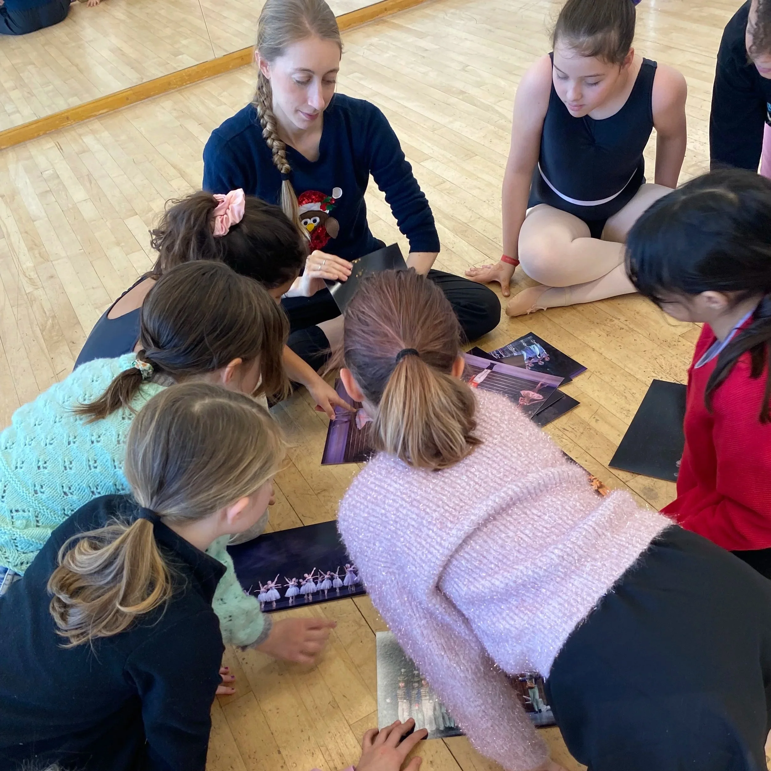 Group of young girls and an adult woman sitting on the wooden floor, looking at photographs of ballet dancers.