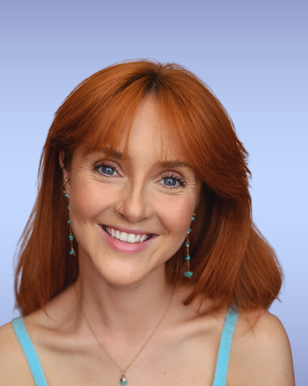 Close-up of a young woman with red hair, blue eyes, and a bright smile, wearing turquoise jewelry and a light blue top against a gradient blue background.